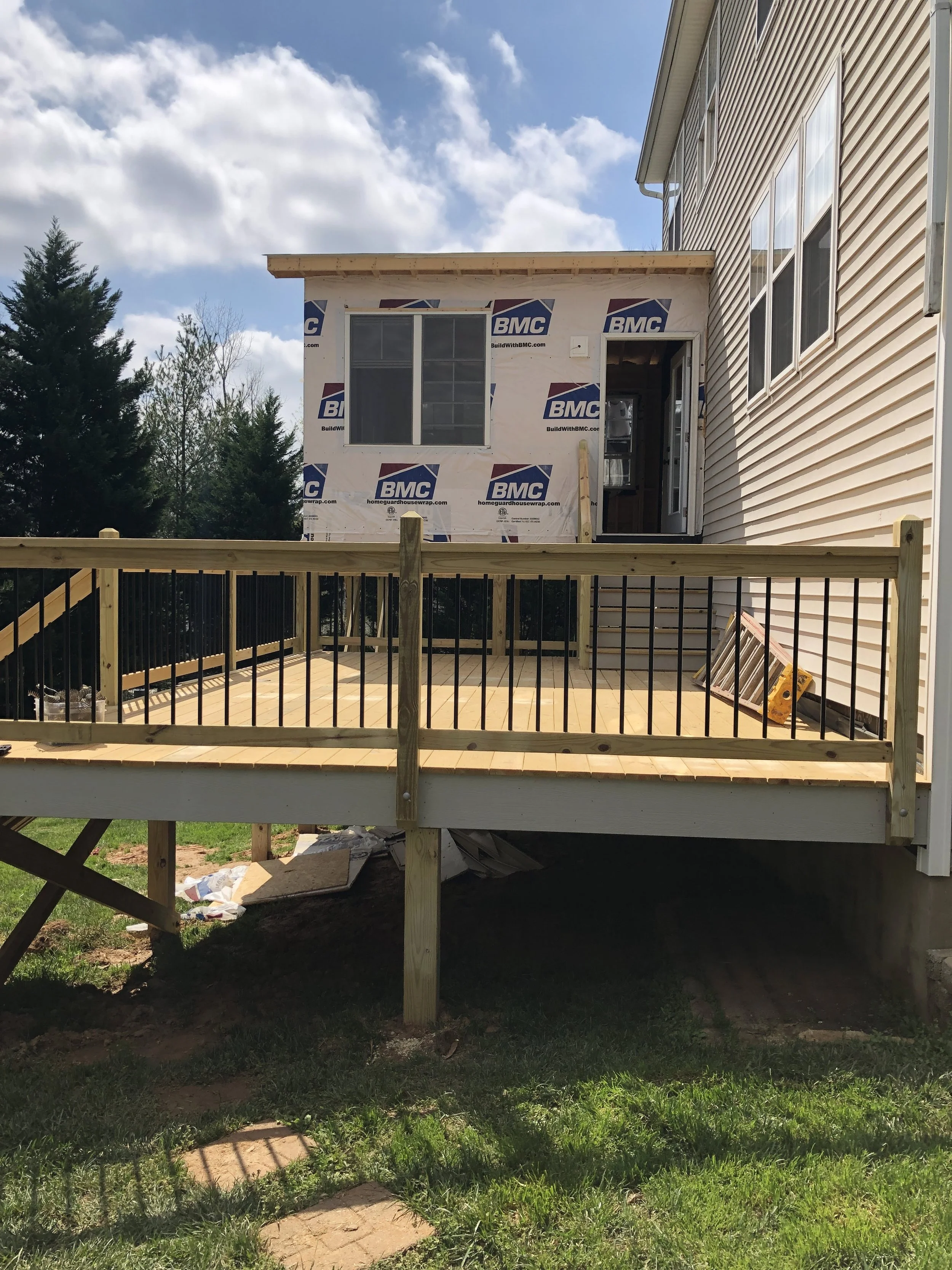 A wooden deck under construction attached to a house with beige vinyl siding. The deck has a railing and a platform with some construction materials underneath. The house has a small extension with wrapped insulation, a window, and a door opening. Th