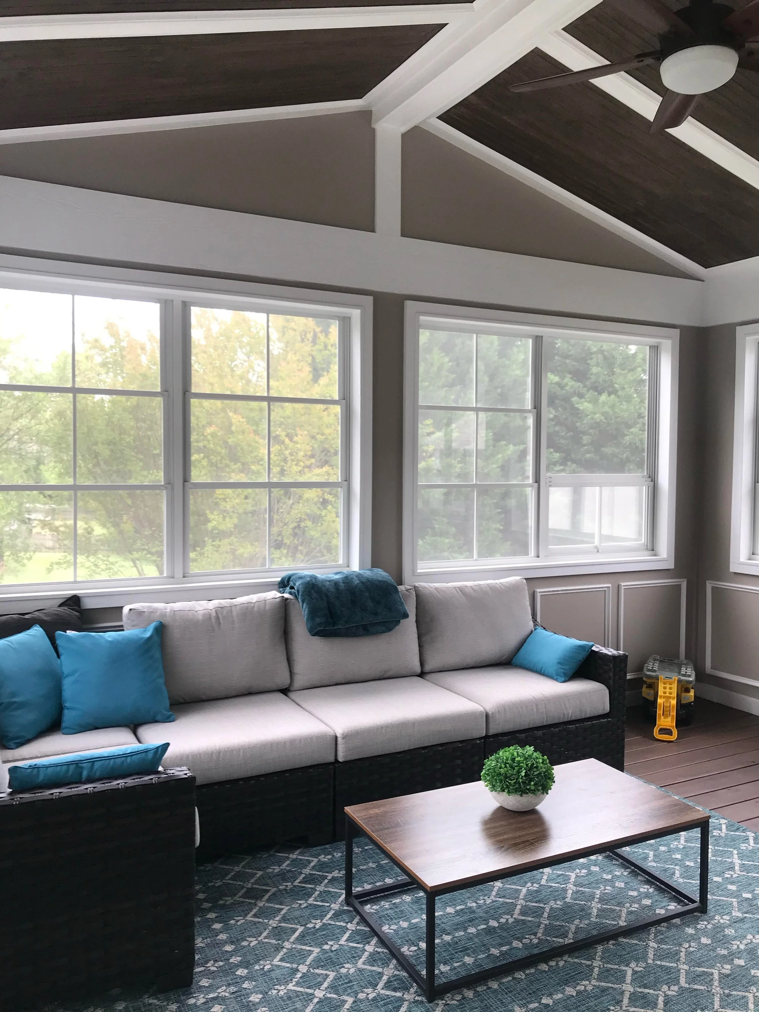 Living room with large windows, beige sectional sofa with blue pillows, a wooden coffee table with a small green plant, a patterned blue rug, and a ceiling fan with dark blades.