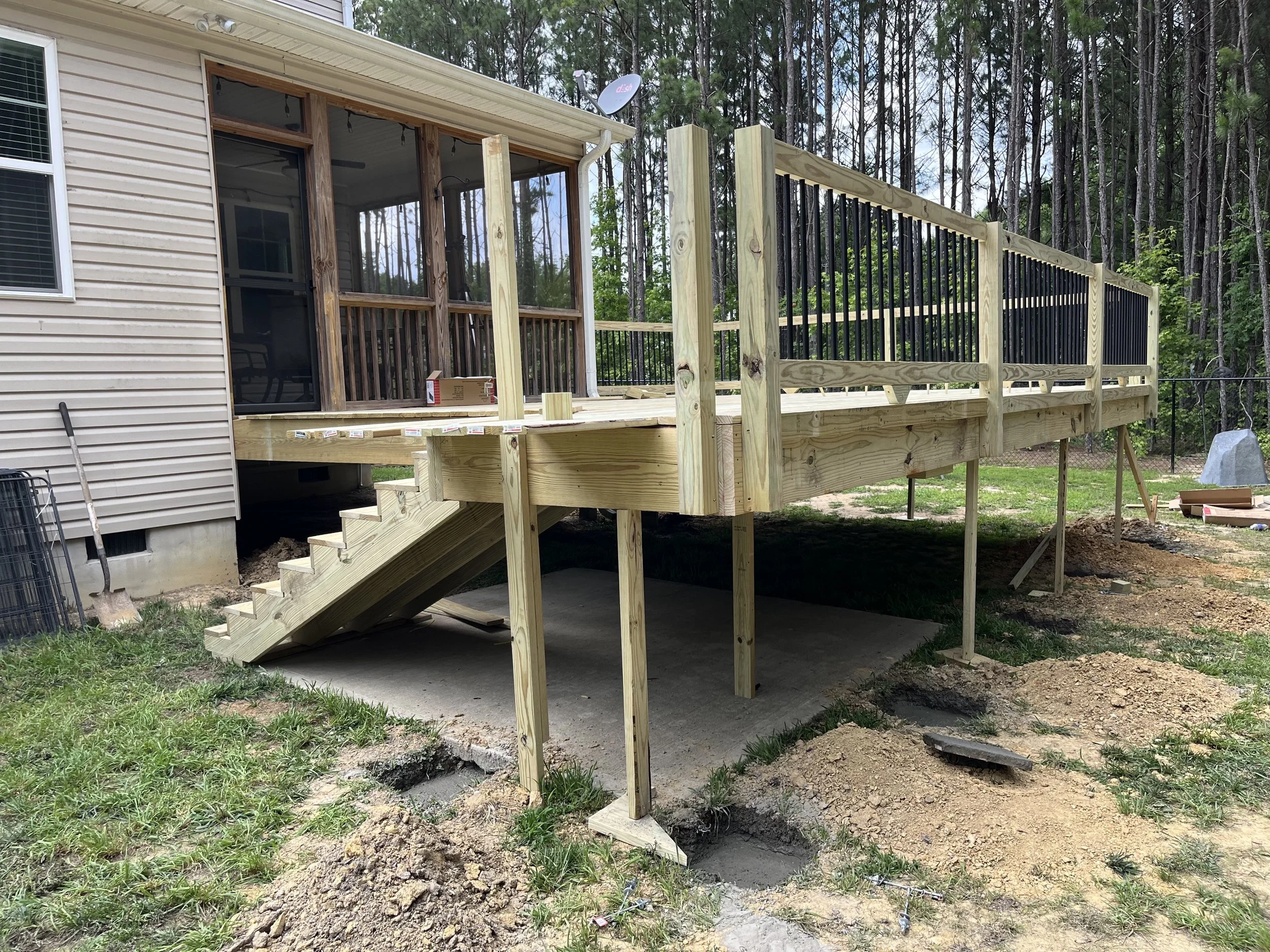 A wooden deck under construction attached to a house, with stairs leading down to the ground, and a safety railing partially installed. There are trees in the background and some construction materials around.