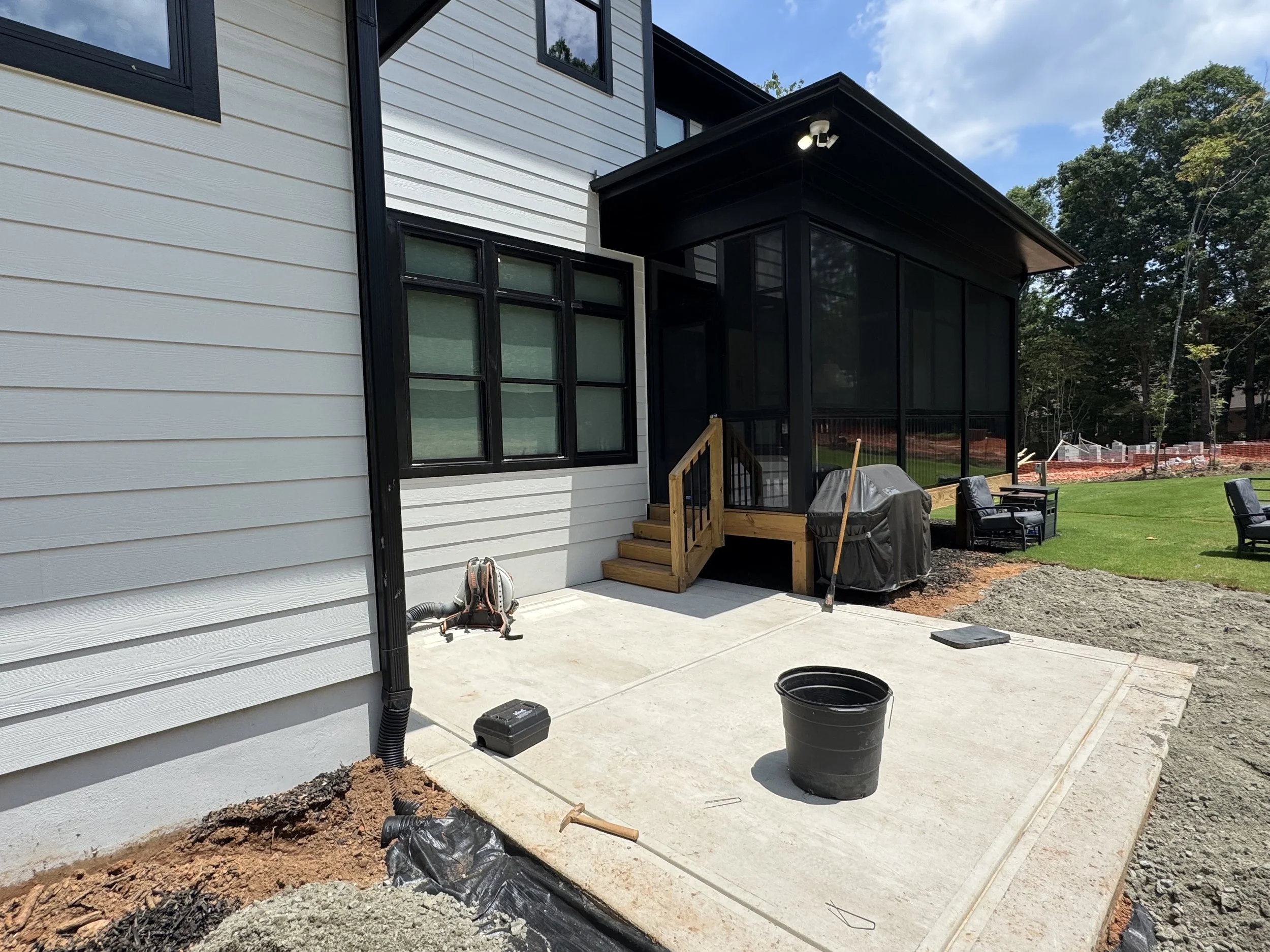 Backyard patio under construction with black framed screened porch, stairs, and outdoor chairs, surrounded by trees and a blue sky.