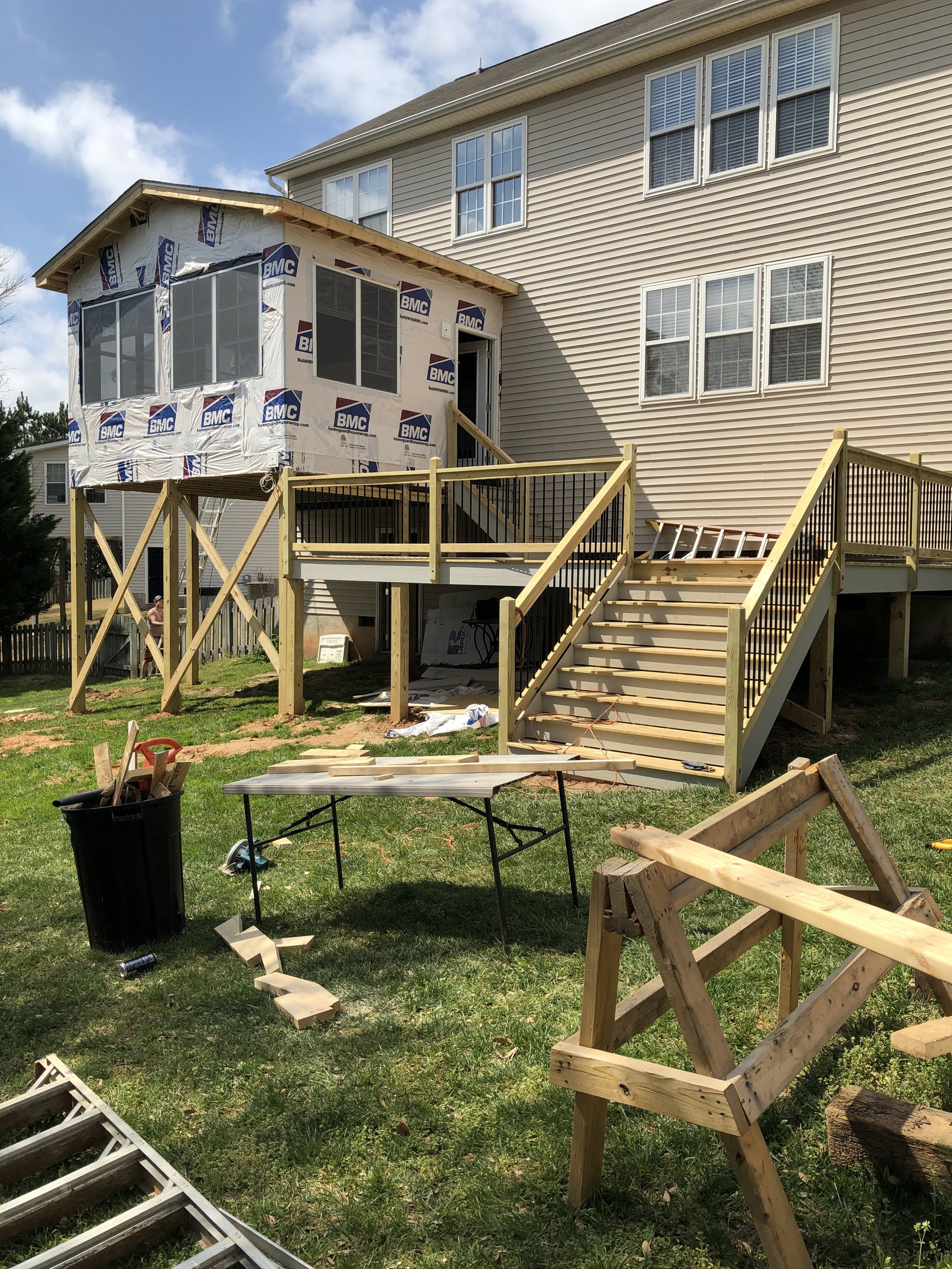 Photo of a house under construction with a raised deck and stairs. The house has beige siding and multiple windows. Construction materials, tools, and wooden framework are visible in the yard, indicating ongoing building work.
