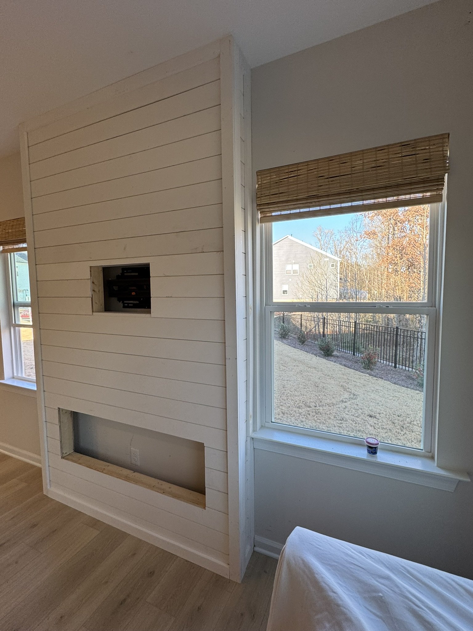 The image shows an interior room with a large window letting in natural light, a white wood-paneled wall with an opening, possibly for a fireplace, and a bed partially visible on the right side with a white sheet. Outside the window, there's a backya
