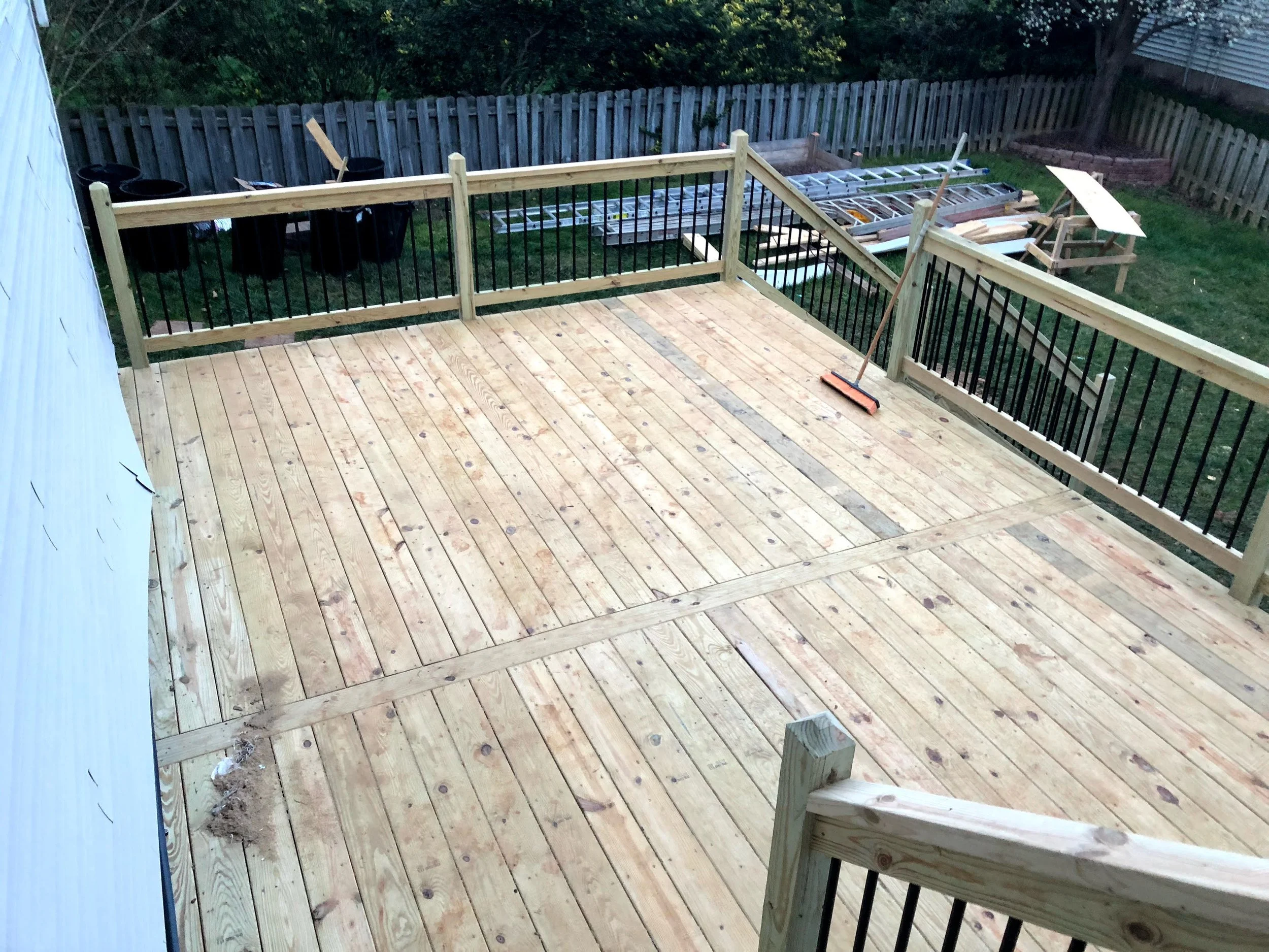 A backyard deck under construction with wooden flooring, railing, and tools, including a broom and construction materials in the background.