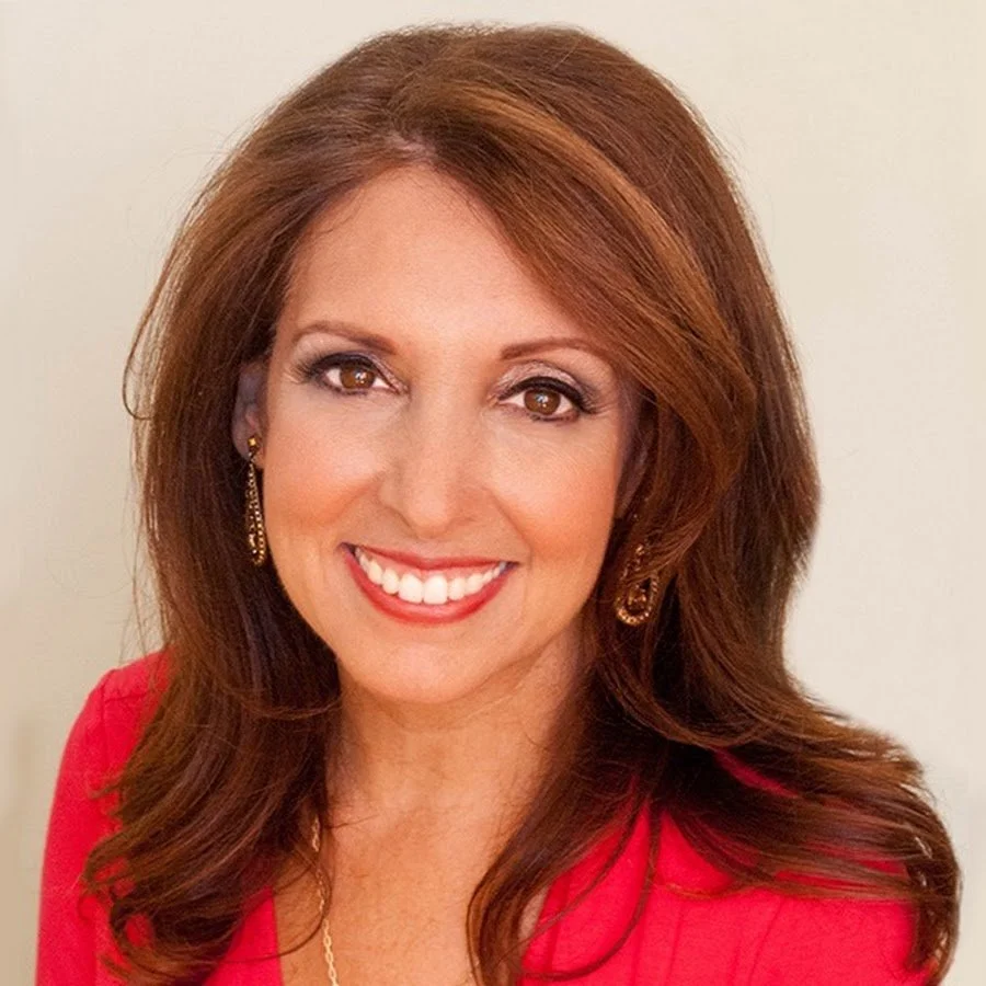 Portrait of a woman with shoulder-length red hair, wearing a red top, gold earrings, and a necklace, smiling against a light background.