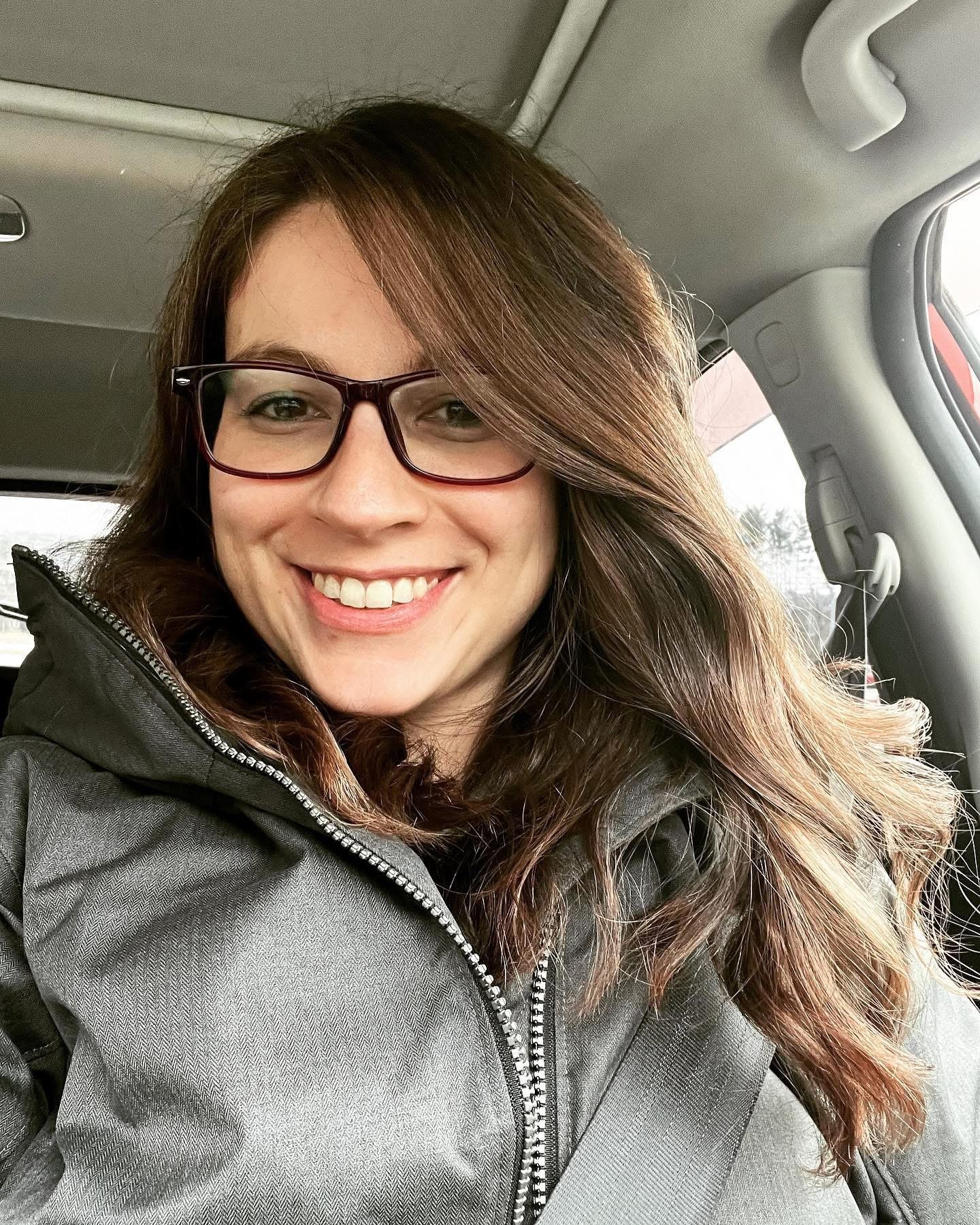 A woman with long light brown hair and glasses smiling in a car, wearing a gray jacket.
