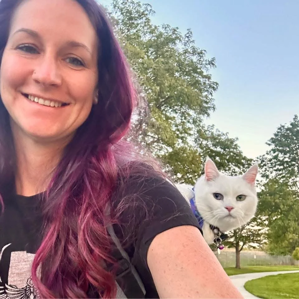 A woman with purple and pink hair smiling at the camera, with a white cat on her shoulder outdoors in a park-like setting with trees and a pathway.
