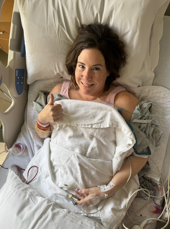 A woman lying in a hospital bed, giving a thumbs-up and smiling, with medical tubes and bracelets on her wrist.