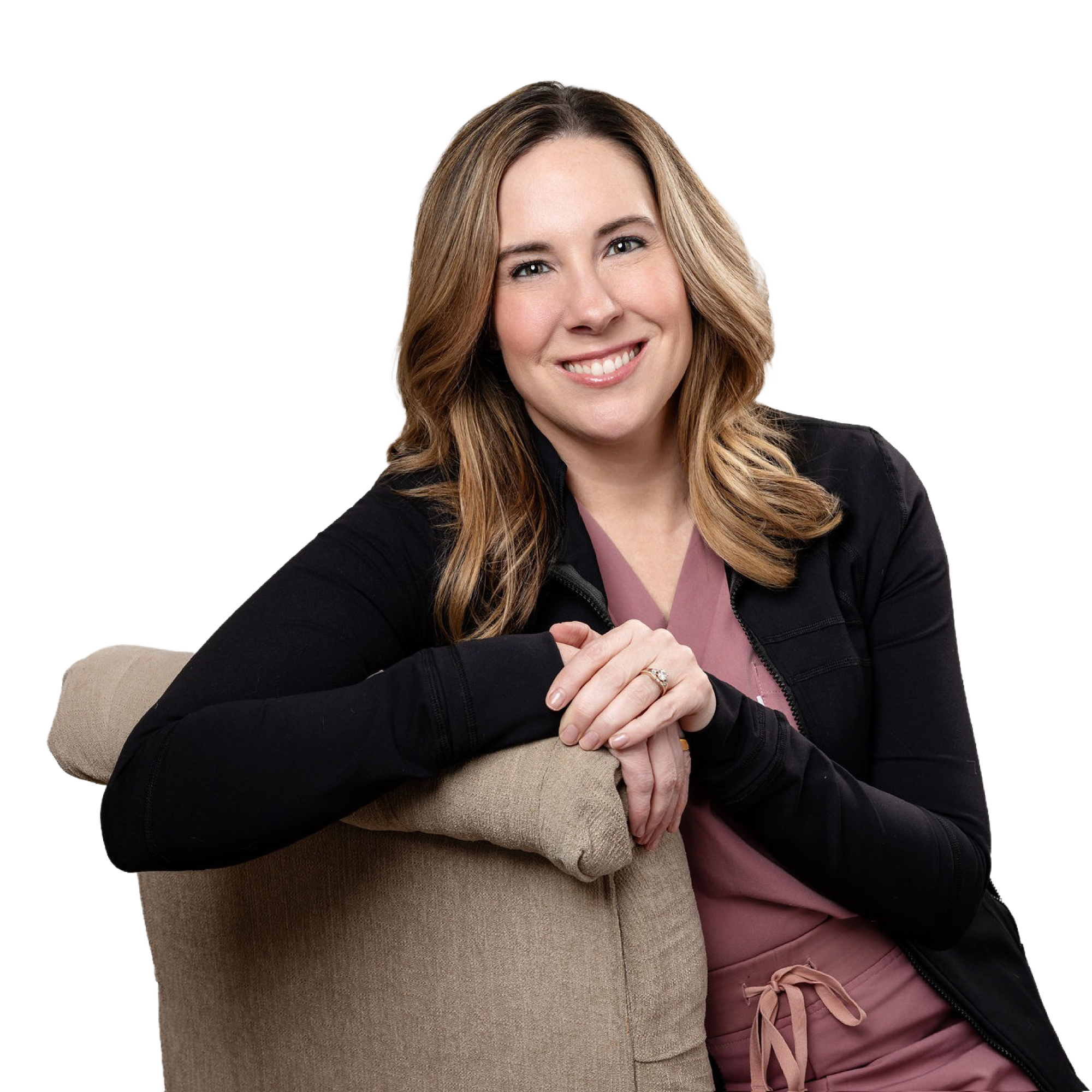 A woman with light brown hair smiling, wearing a black jacket over a pink top, sitting with an arm around a person wearing a beige coat. The sweater's arm is visible, and the person is wearing a ring. The background is plain and dark.