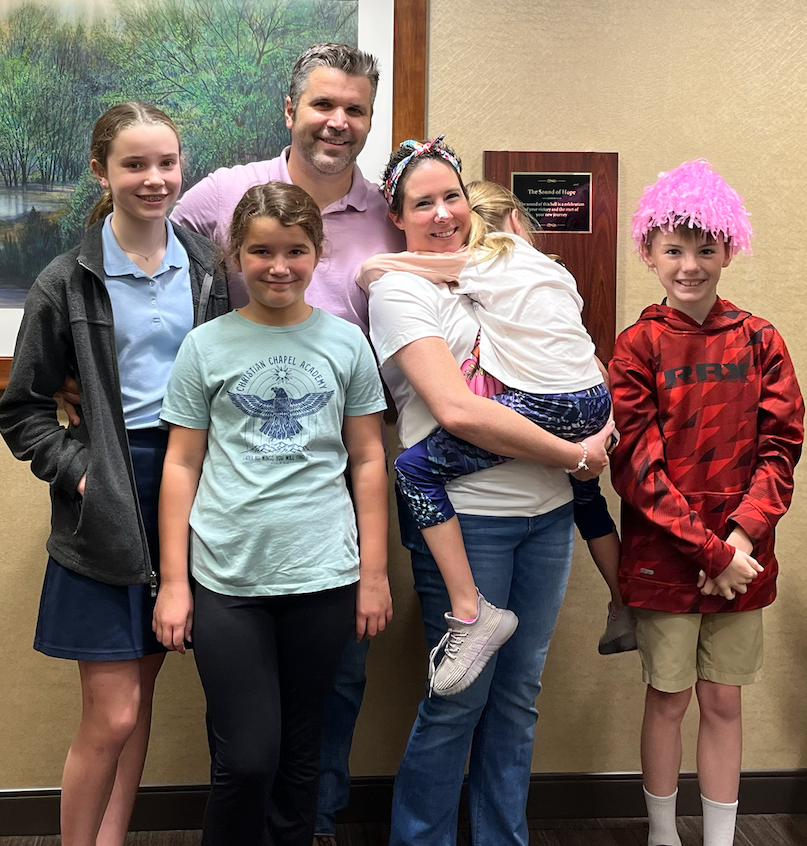 A family of five posing indoors in front of a wall with a framed picture, smiling. The mother is holding a young girl with a pink wig, and other children stand with them.