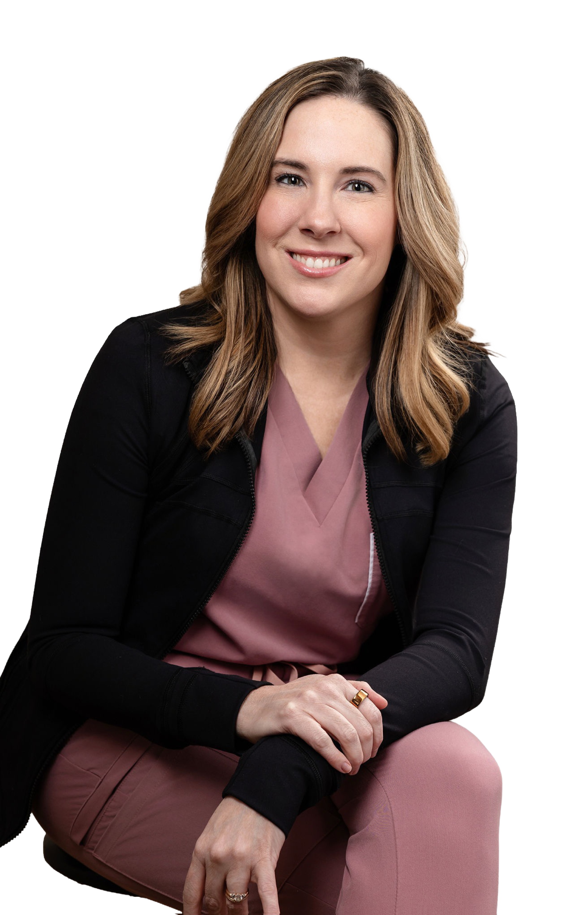 A woman with long, wavy blonde hair and a bright smile, wearing a black jacket over a pink top and pants, sitting with her left arm resting on her lap and her right hand on her knee, against a white background.