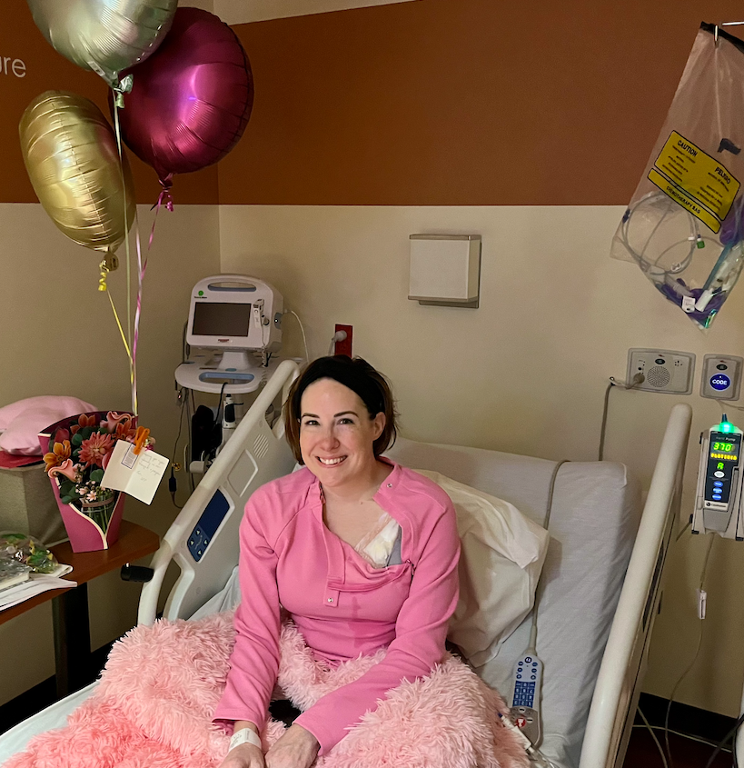 Woman sitting in hospital bed wearing pink gown, smiling, with balloons, flowers, and hospital equipment around her.