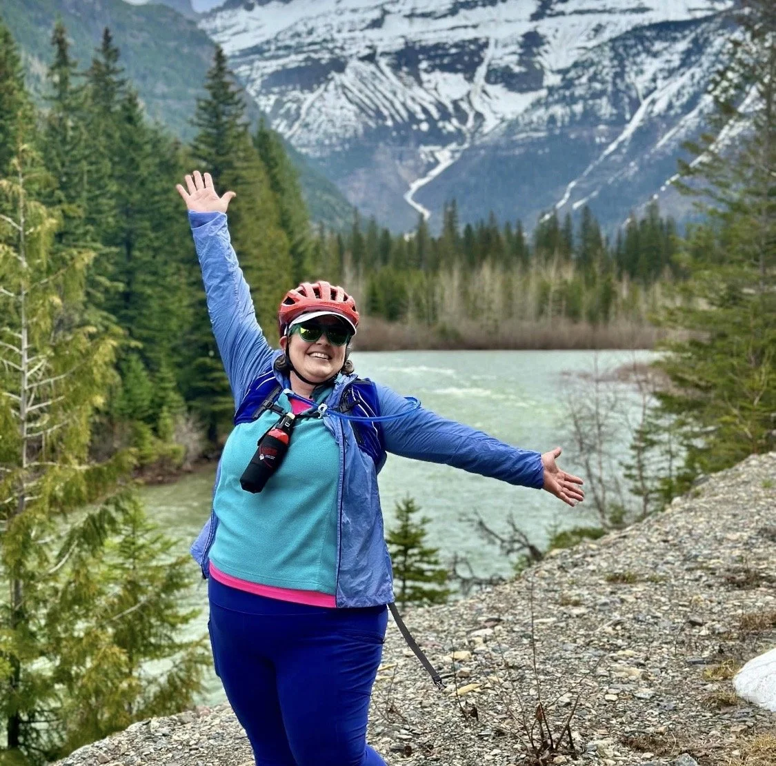 A woman in outdoor gear, wearing sunglasses and a helmet, standing near a river with her arms raised in a joyful pose, surrounded by a forest and snow-capped mountains.
