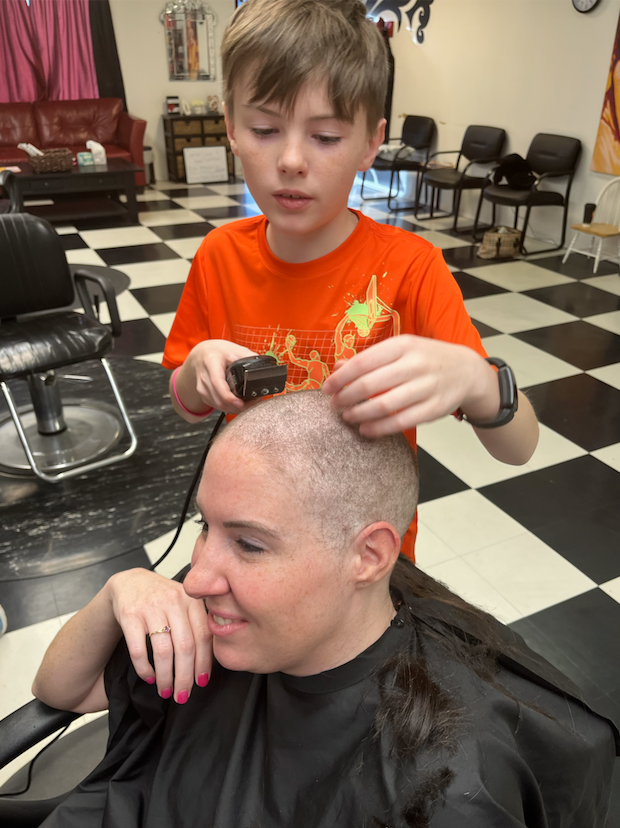 A young boy with a camera is photographing a woman with very short or shaved hair, who is sitting in a salon chair. The salon has a checkered black and white floor, black chairs, and some furniture and decorations in the background.
