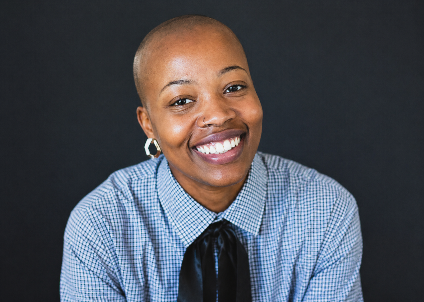 A smiling woman with a shaved head, wearing a blue and white checkered shirt and a black tie, against a dark background.