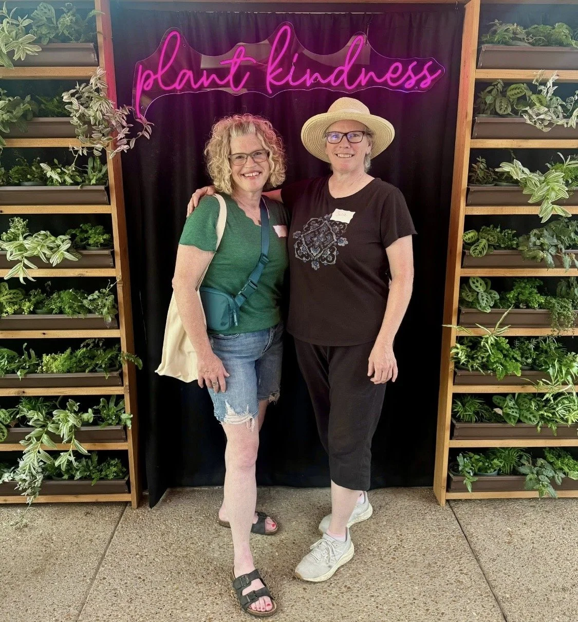 Two women standing in front of a black backdrop with pink neon sign that says "plant kindness". They are smiling, one wearing a green shirt, denim shorts, and sandals, the other wearing a black shirt, black pants, a straw hat, and sneakers. They are surrounded by shelves filled with green plants.