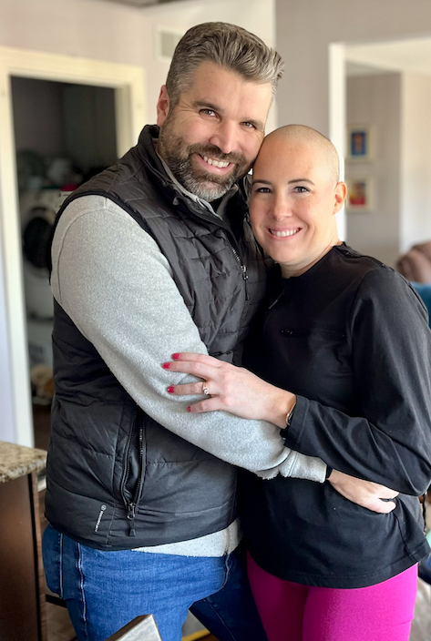 A smiling couple, a man with a beard and a woman with a shaved head, hugging in a home interior.