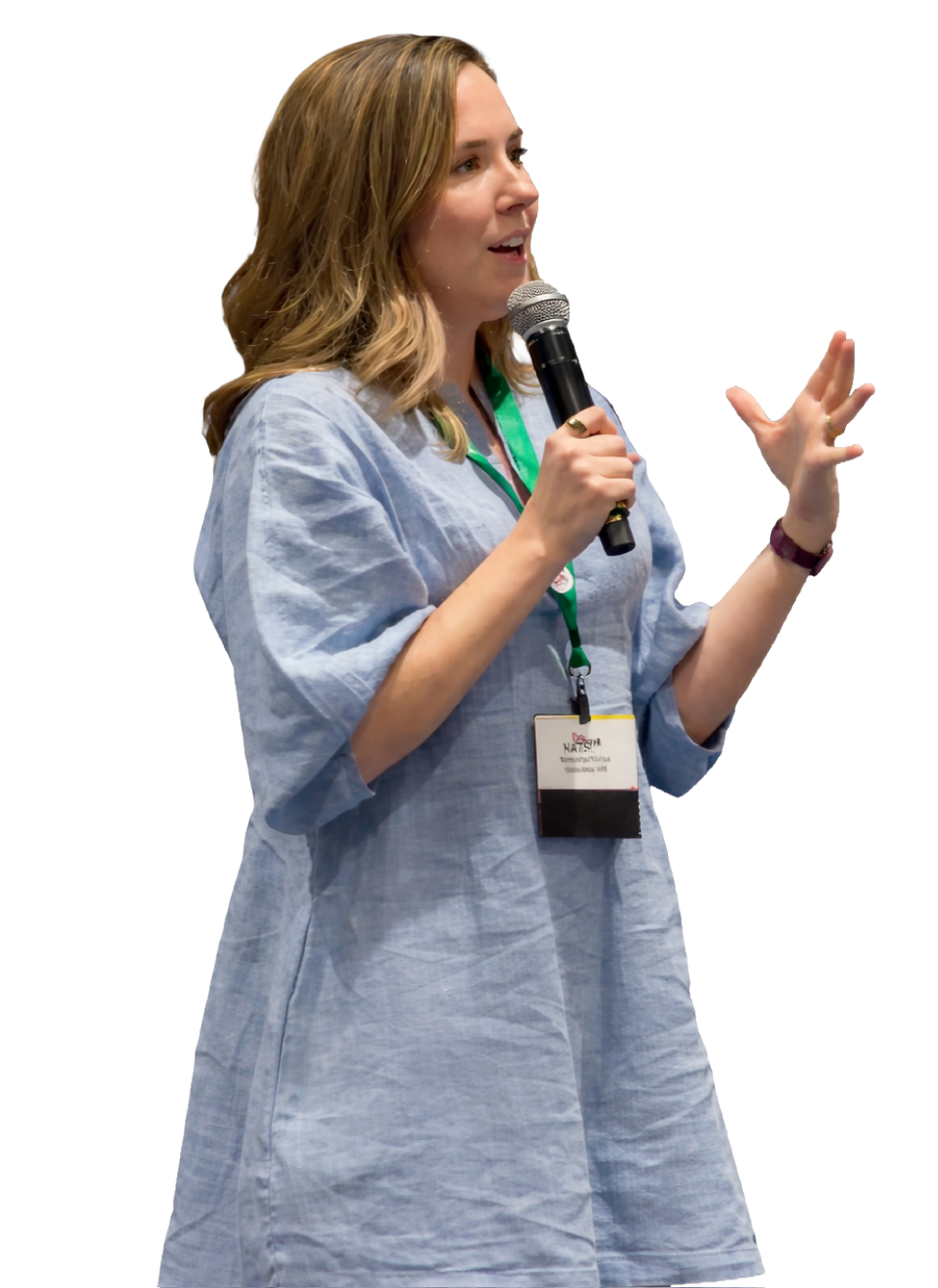 A woman with shoulder-length light brown hair, wearing a light blue dress and a conference badge, is speaking into a microphone and gesturing with her right hand.