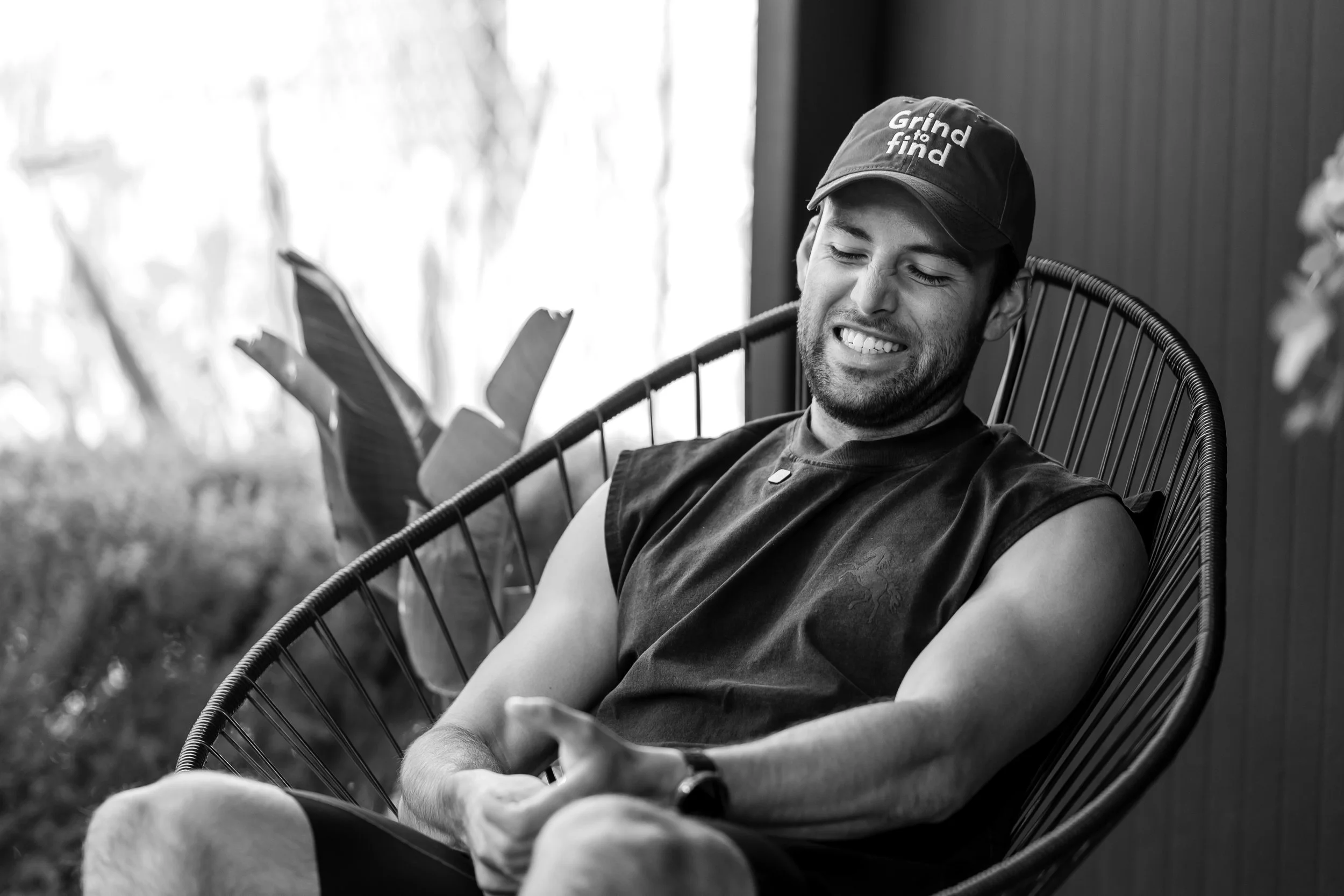 Joven sonriendo mientras mira su teléfono, sentado en una silla de mimbre, con gorra que dice 'Grind to find', en un entorno interior con luz natural.