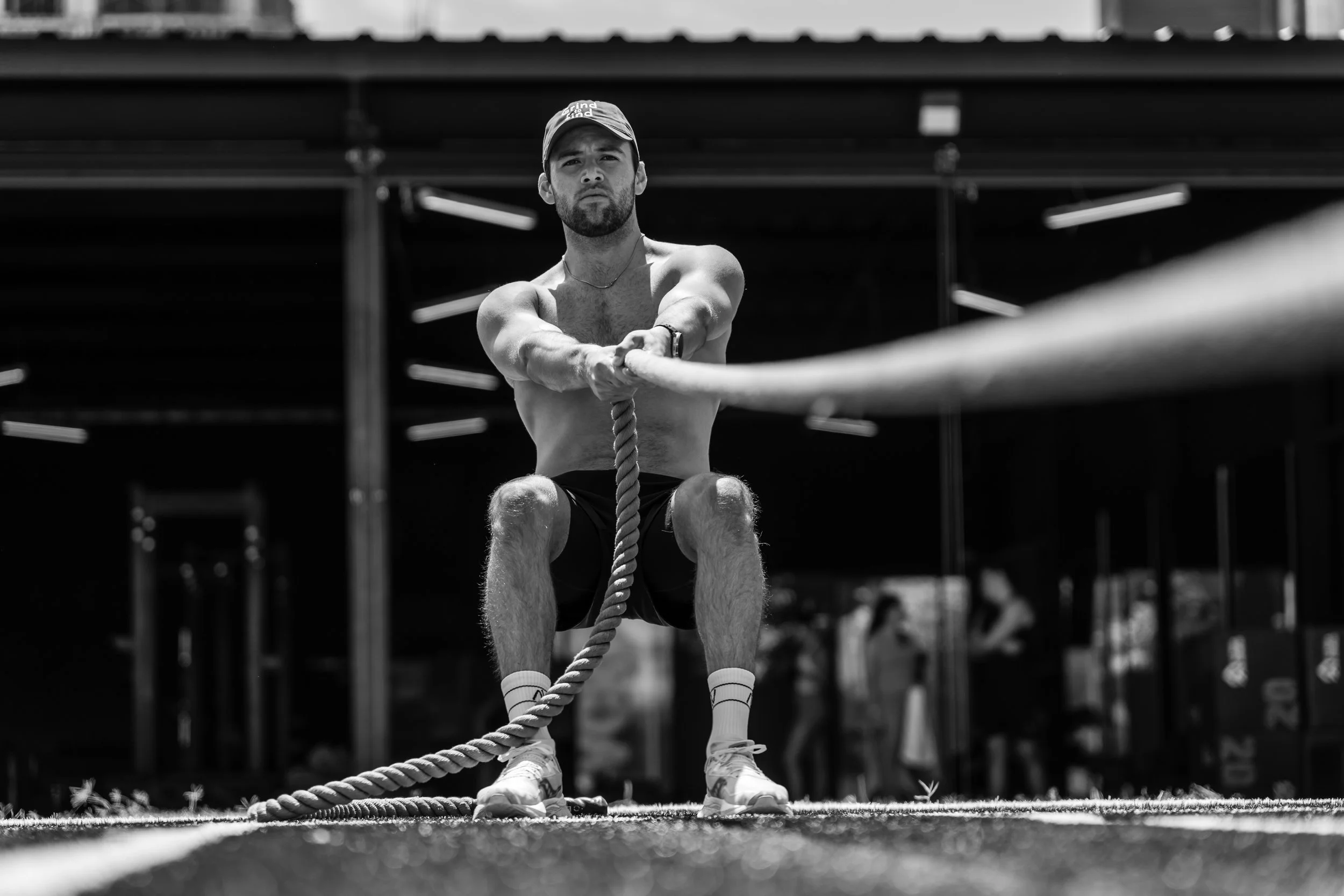 Hombre sin camisa, con gorra y calcetas blancas, haciendo ejercicio de tracción en una cuerda en un gimnasio al aire libre, en color negro y blanco.