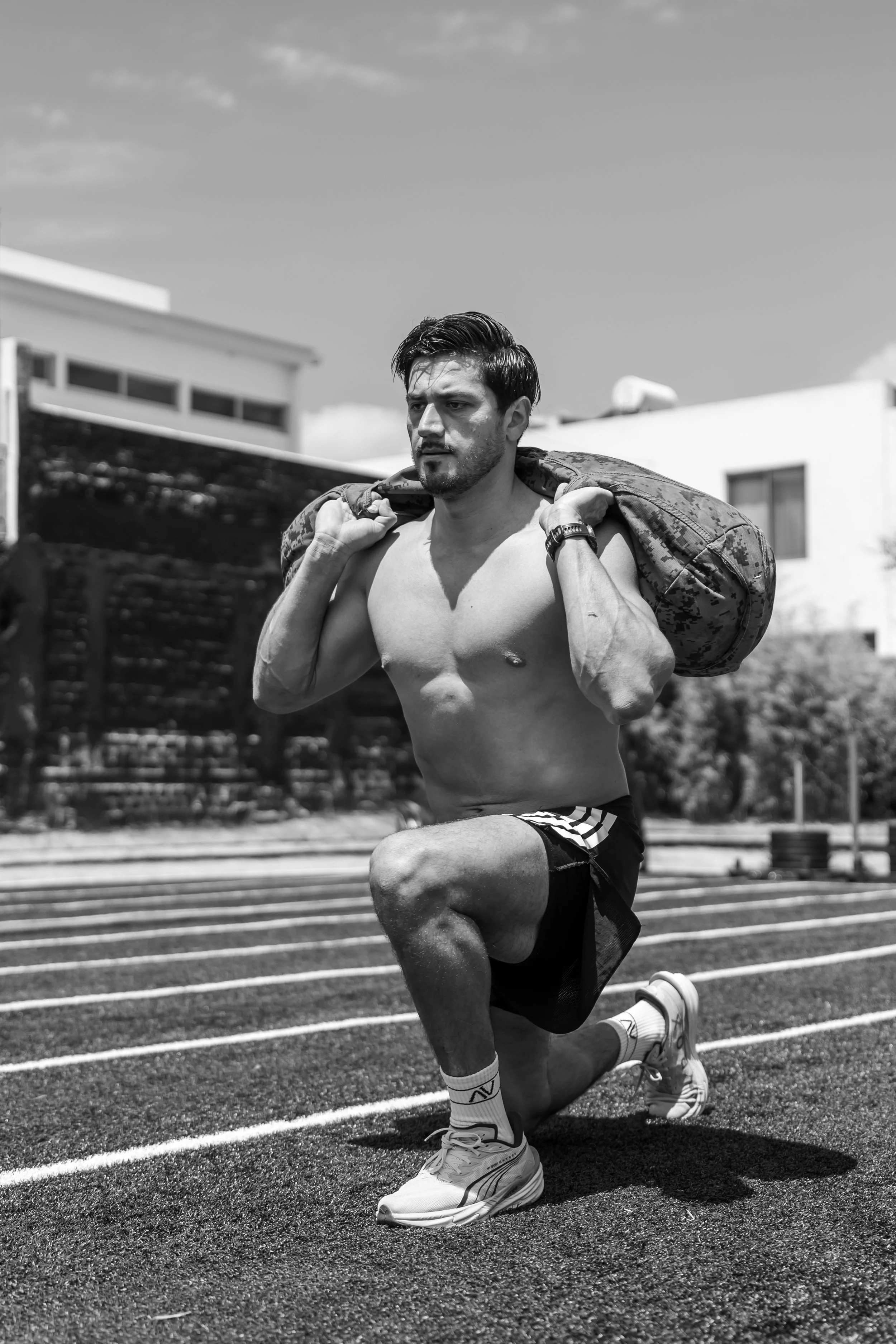 Hombre sin camiseta, en cuclillas en una pista de atletismo, cargando una mochila en los hombros, con el fondo de edificios y un cielo despejado.