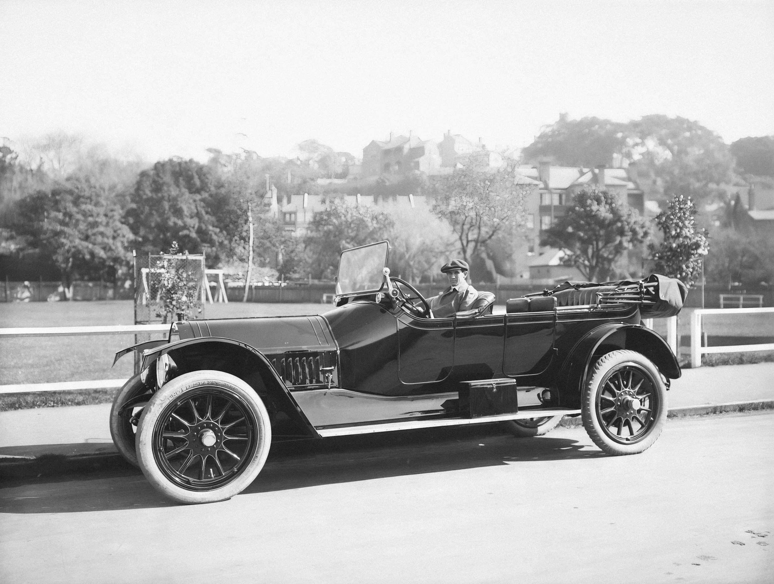 A vintage automobile with a man sitting in the driver’s seat, parked on the side of a street with trees, houses, and hills in the background.