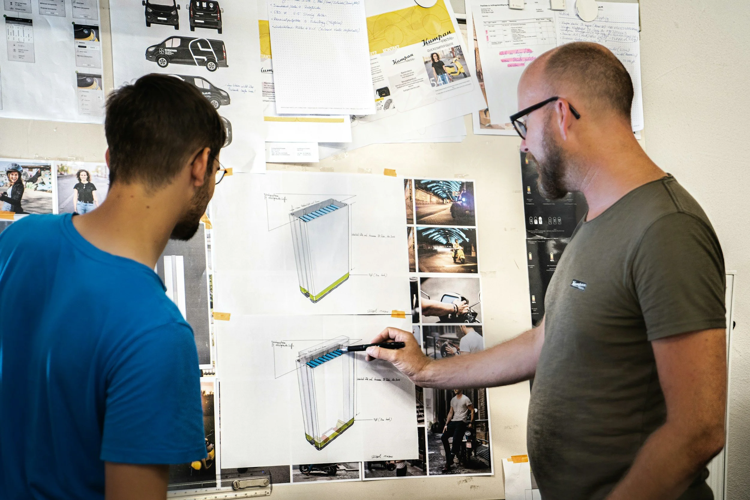 Two men are discussing design plans pinned on a wall. The wall has sketches, photos, and notes, including vehicle illustrations and nighttime street scenes.