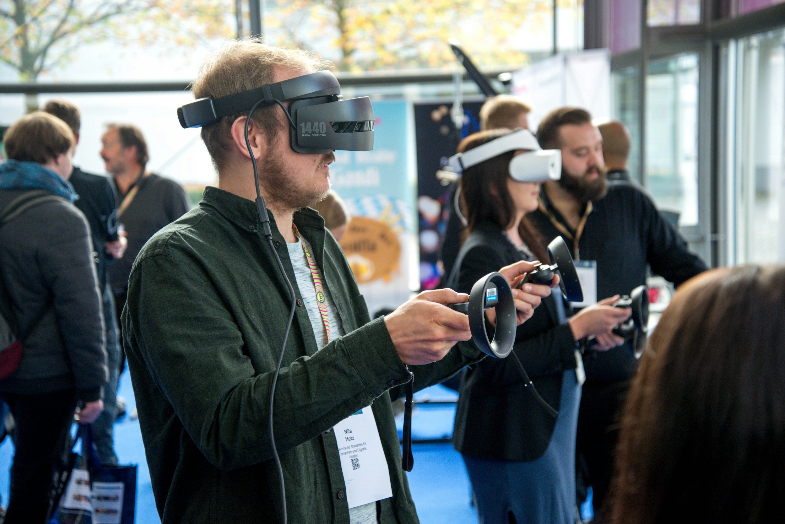 Man wearing a VR headset and holding VR controllers at a technology event, with other people using VR headsets in the background.