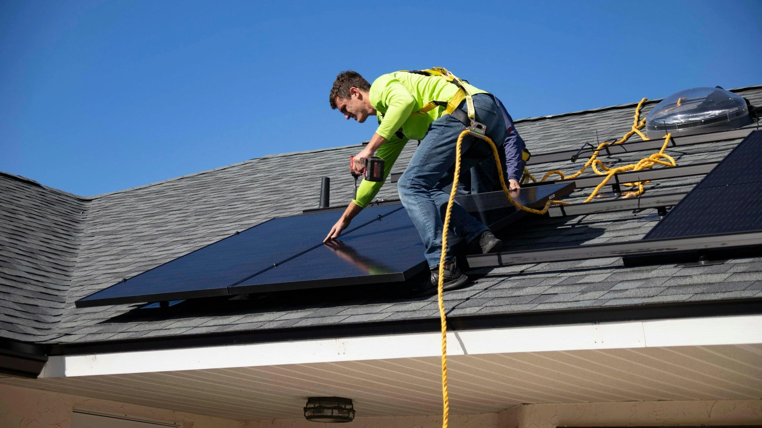 A man in a neon yellow shirt installing solar panels on a home's roof under a clear blue sky.