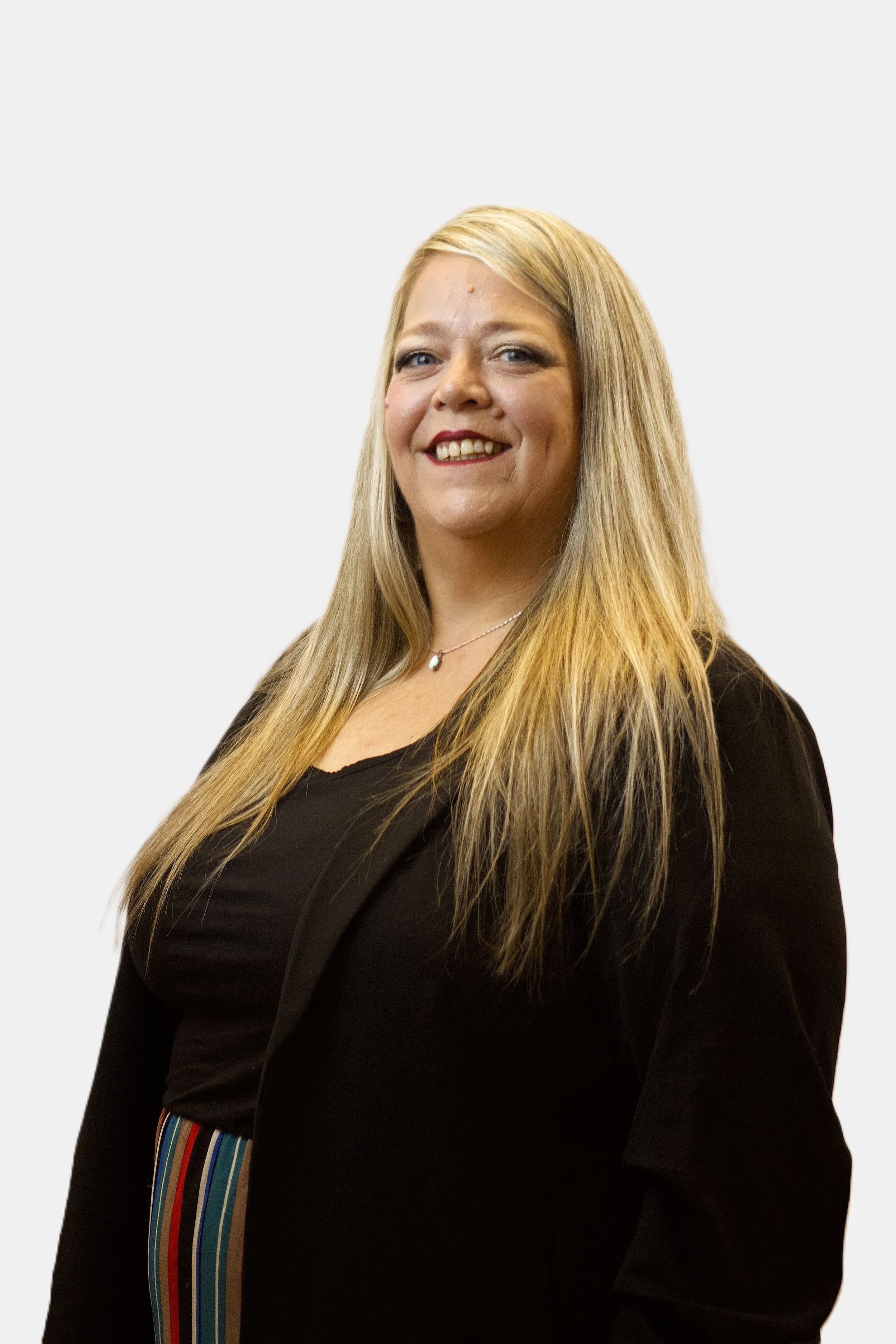 Portrait of a smiling woman with long blonde hair, wearing a black top and a striped belt, standing against a plain light grey background.