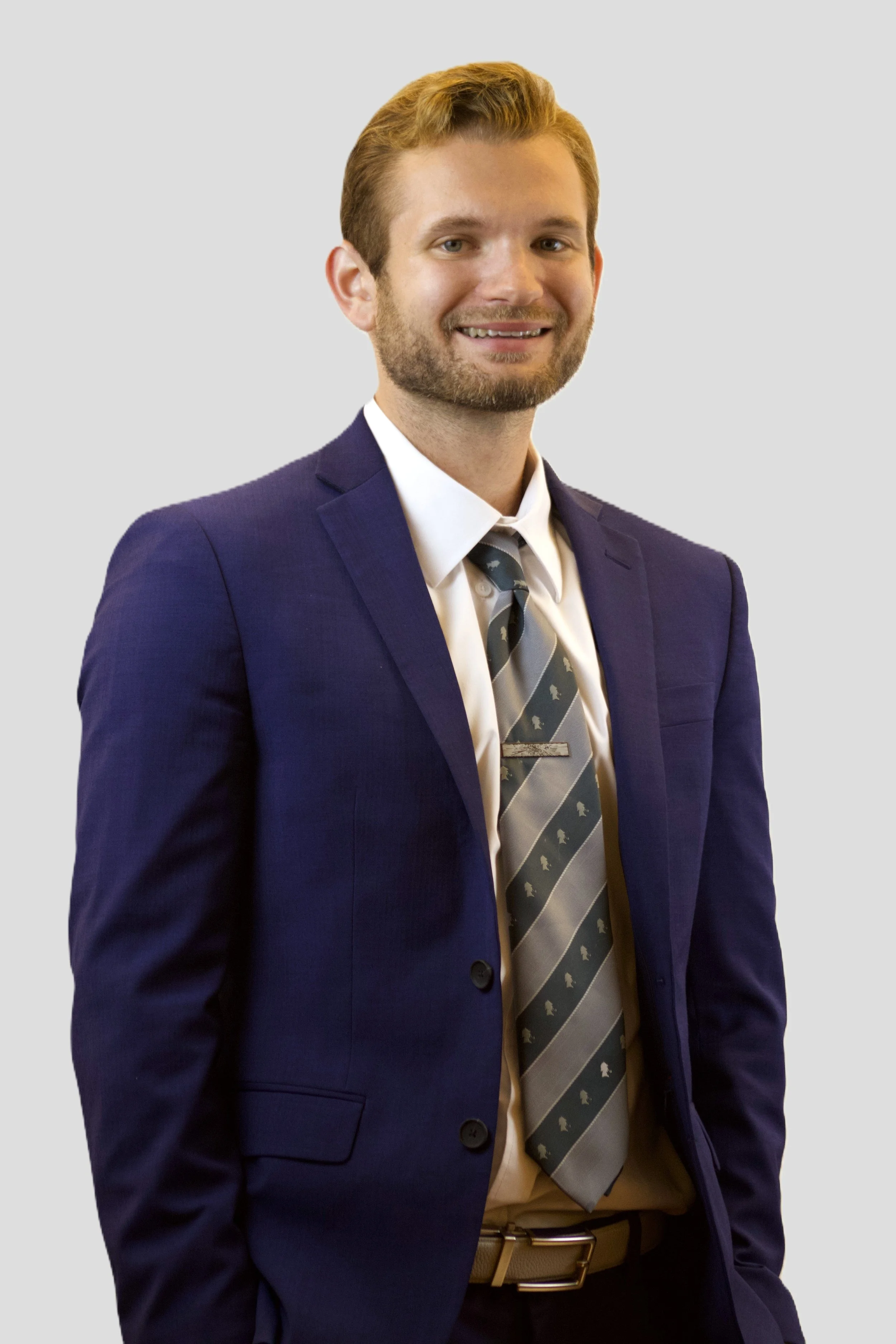 A man in a blue suit, white shirt, and patterned tie standing against a plain light gray background.
