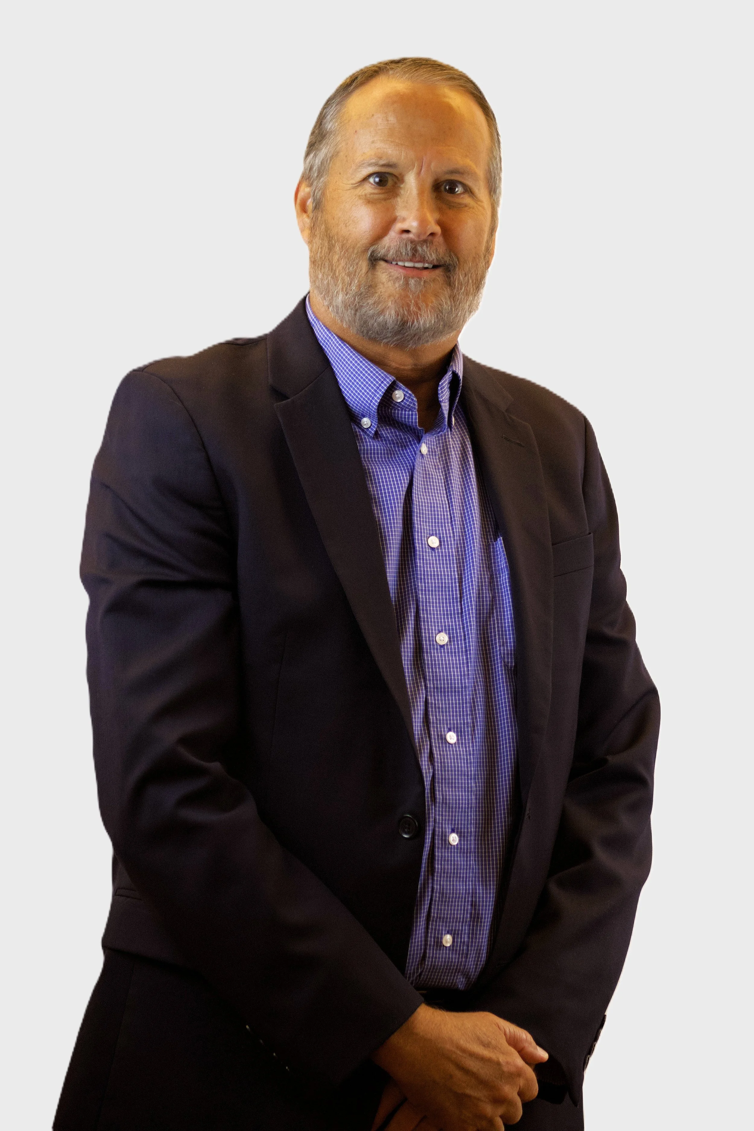 A man with a beard and mustache dressed in a dark suit jacket over a blue checkered shirt, standing with his arms crossed against a plain white background.