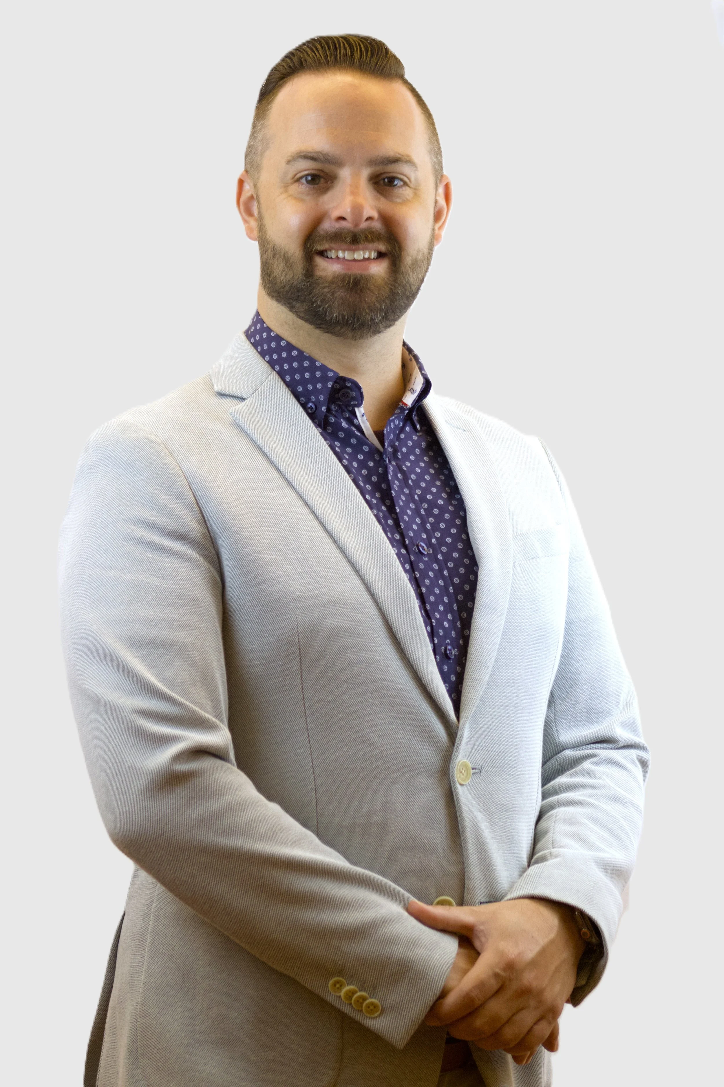 A man in a light-colored blazer and a blue polka-dot shirt, smiling with his arms crossed, against a plain light background.