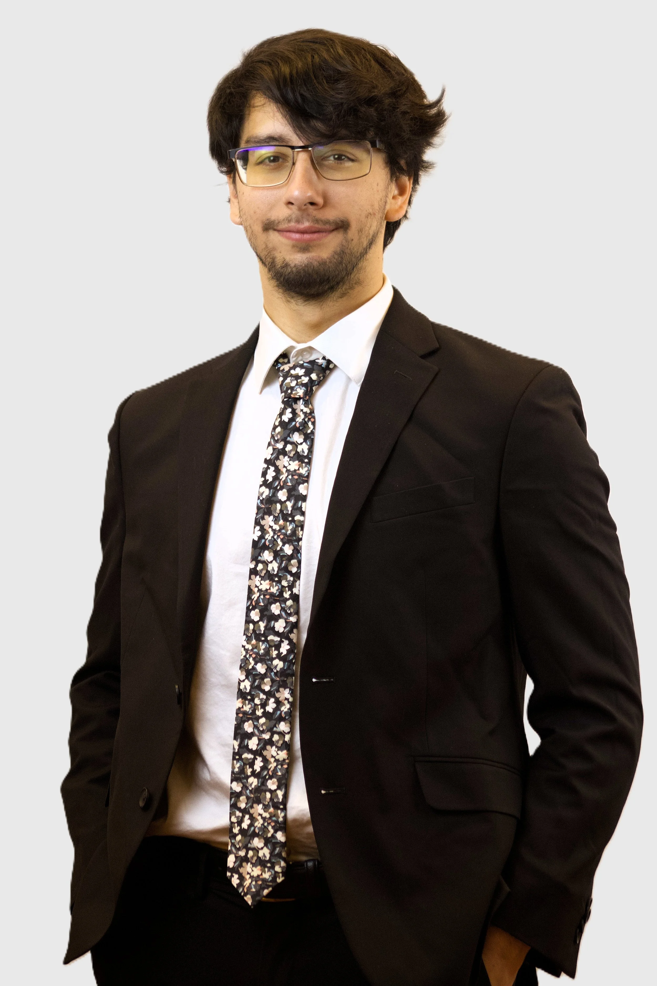A young man with dark, wavy hair and glasses wearing a black suit, white shirt, and a floral tie, standing against a plain light gray background.