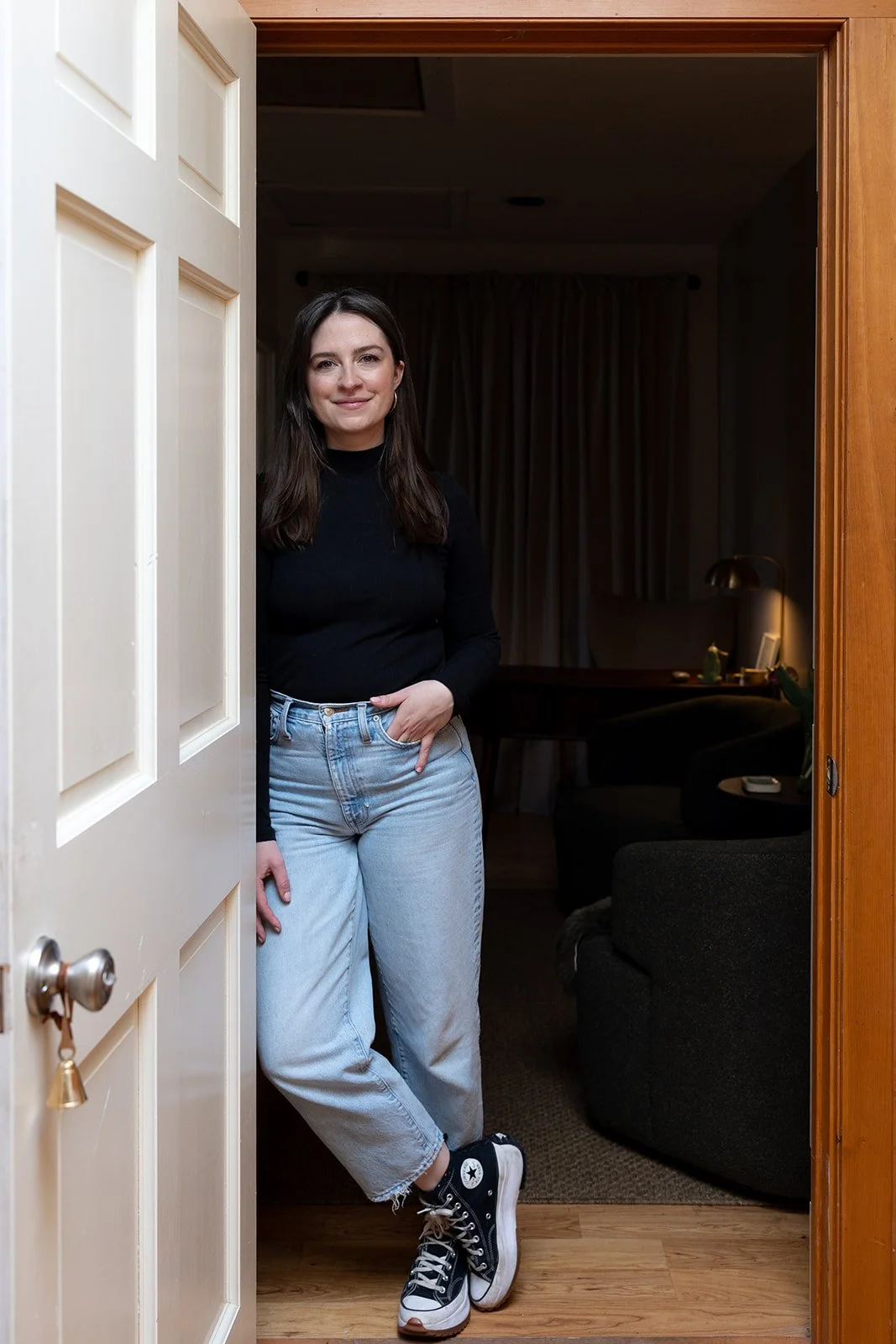 A young woman with shoulder-length dark hair, wearing a black long-sleeve top, light-wash jeans, and black high-top sneakers, stands in a doorway inside a home with her hands in her pockets, smiling at the camera.