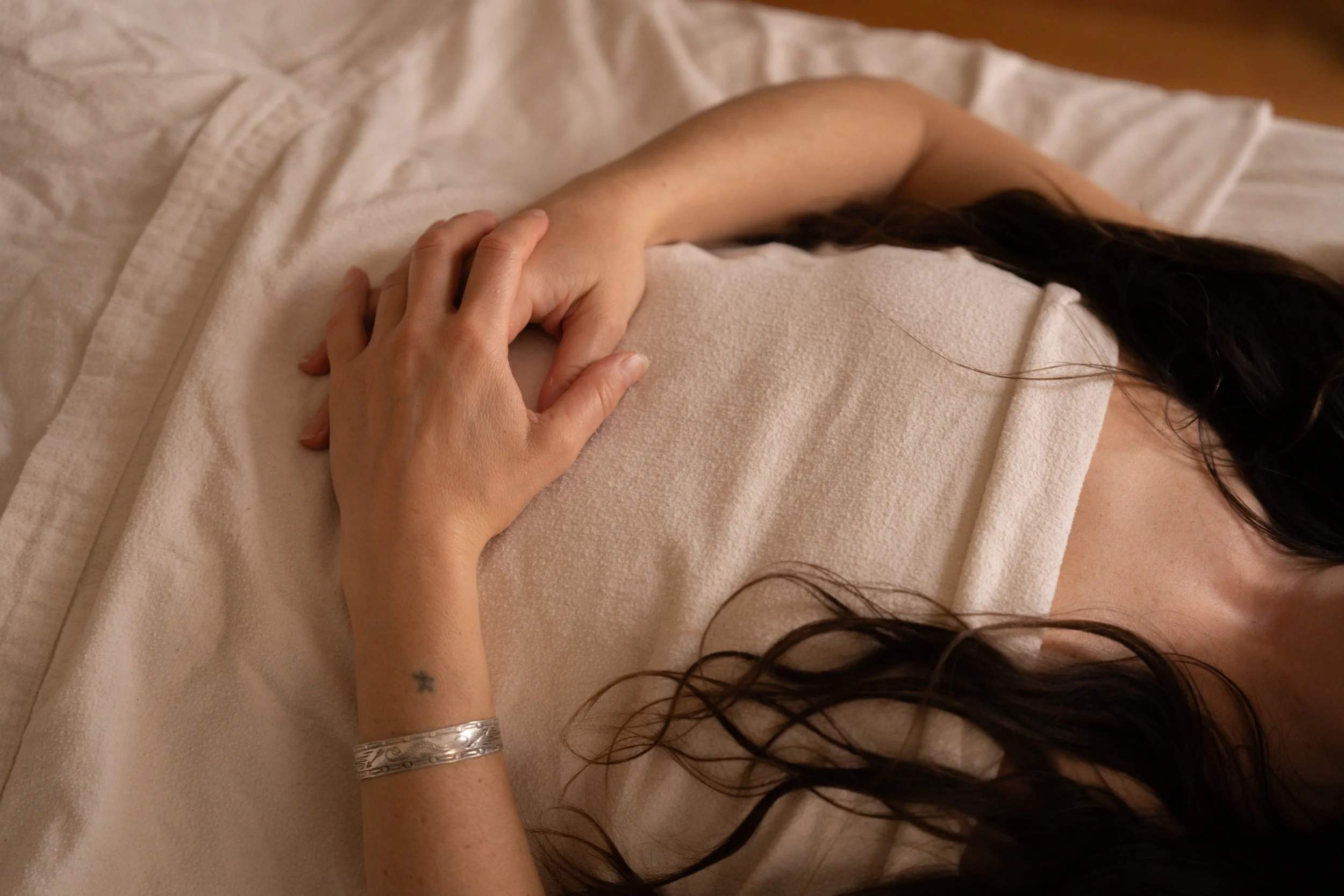 A person with long dark hair lying on a bed in a beige robe, touching their arm, with a silver bracelet and a small star tattoo visible on their wrist.