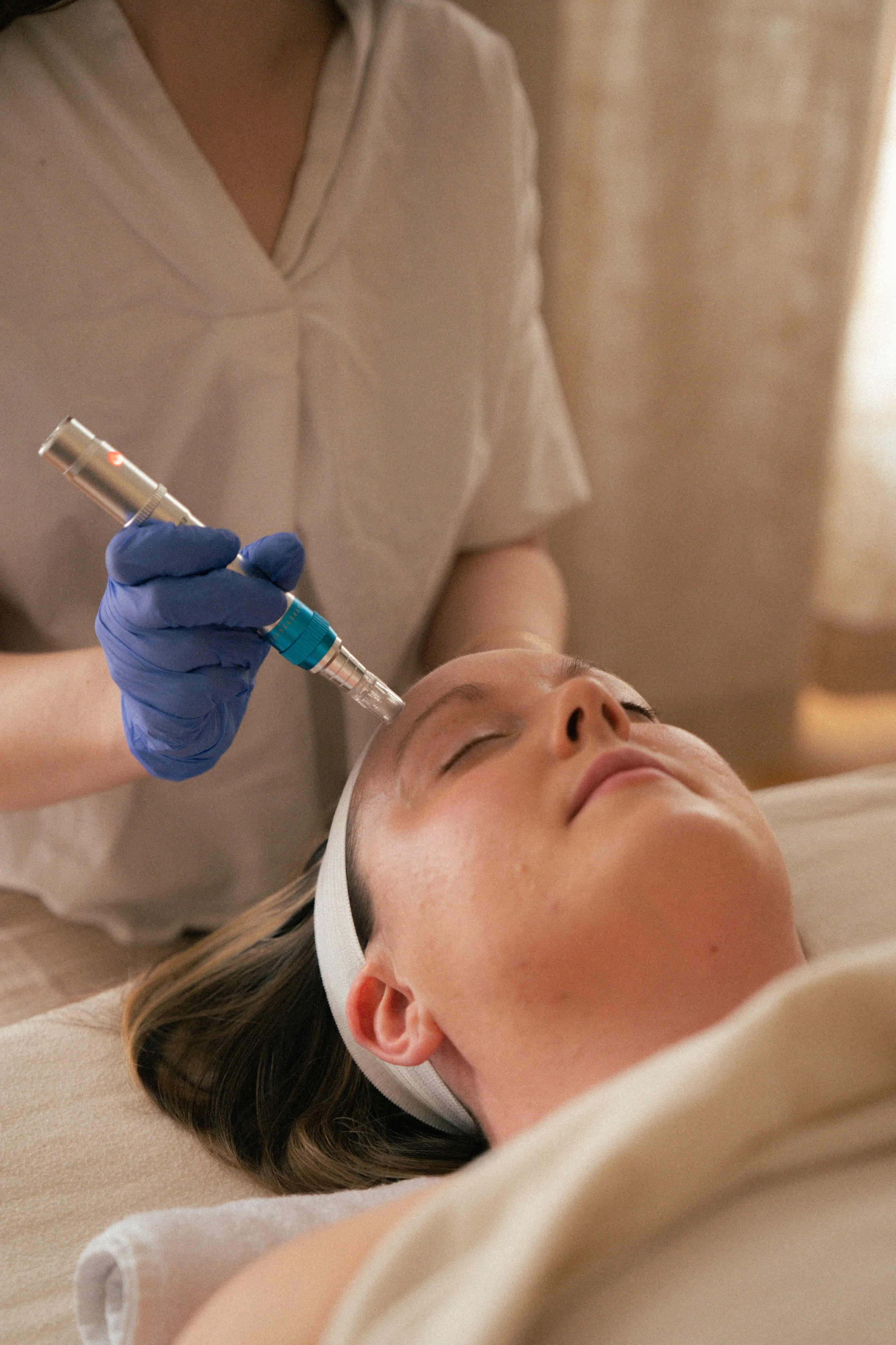 A person lying on their back with eyes closed, receiving a cosmetic or medical treatment on their face from a practitioner wearing blue gloves, in a clinical setting.