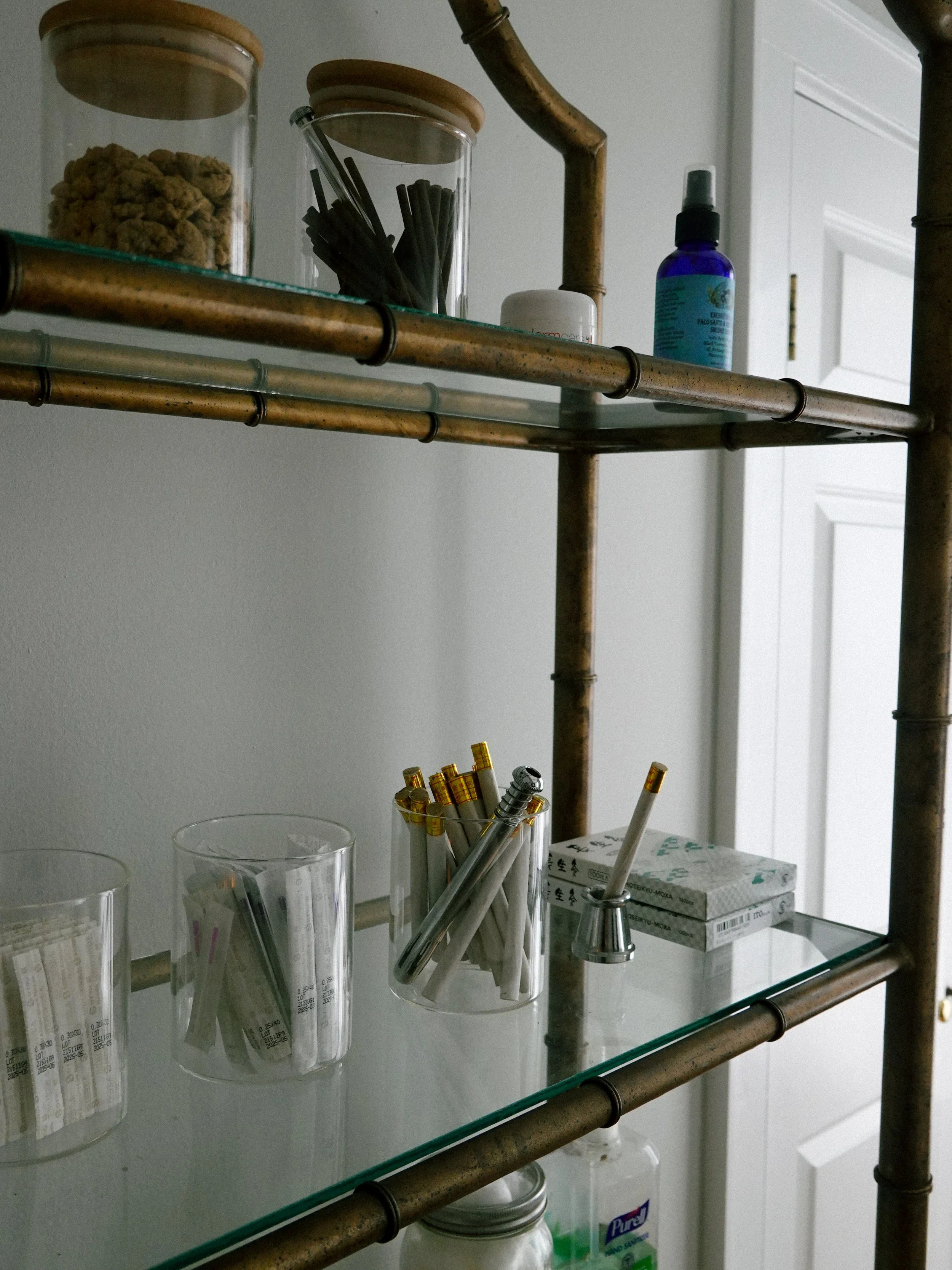 A glass shelf with various items including clear measuring cups, a glass with pens and pencils, a box with Asian characters, and a spray bottle, against a white wall and door.