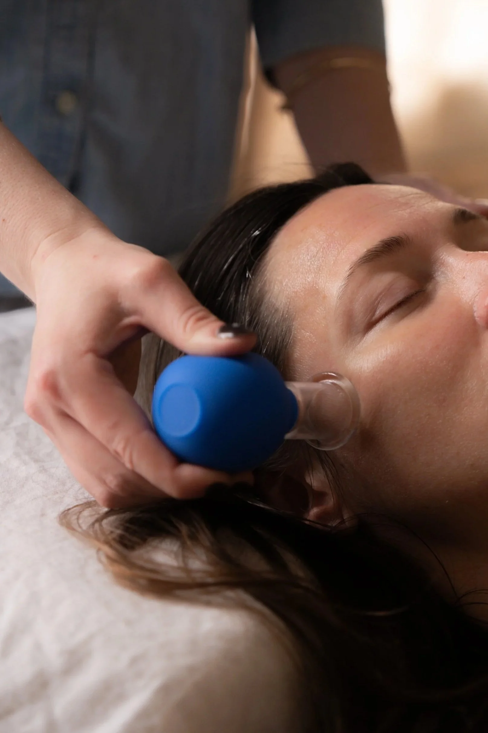 A woman with dark hair receiving a scalp treatment with a blue tool from a practitioner wearing gray clothing.