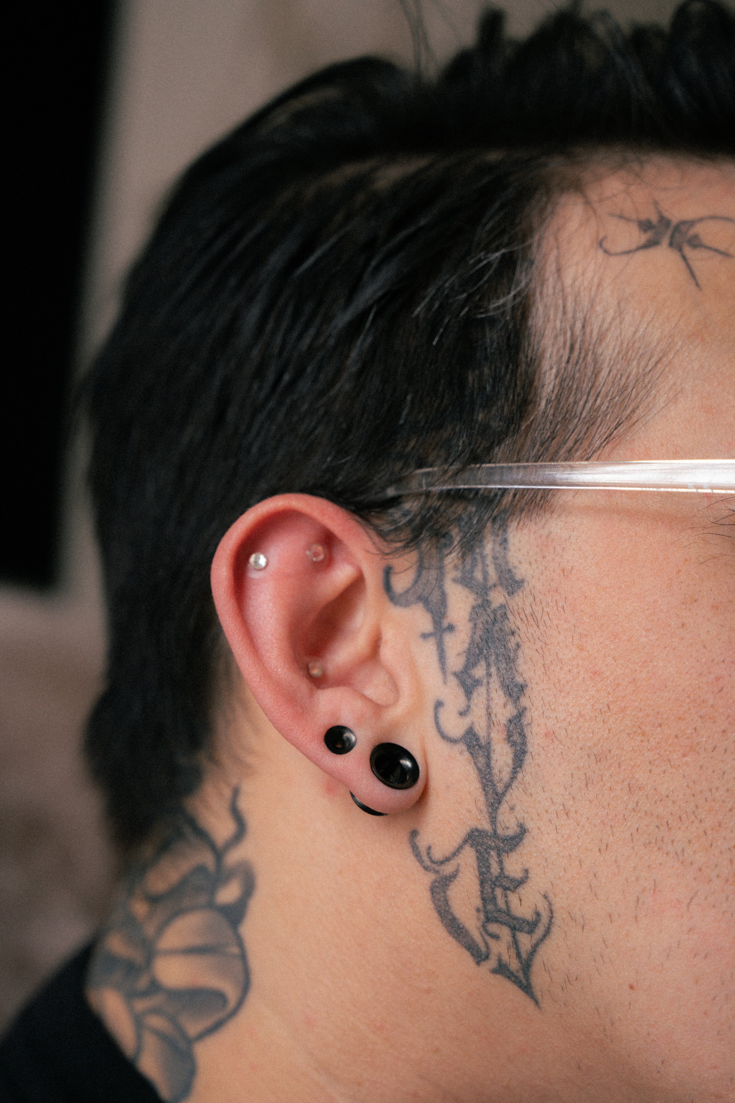 Close-up of a person's side face showing multiple tattoos and earrings, with black hair styled short and layered, wearing clear glasses.