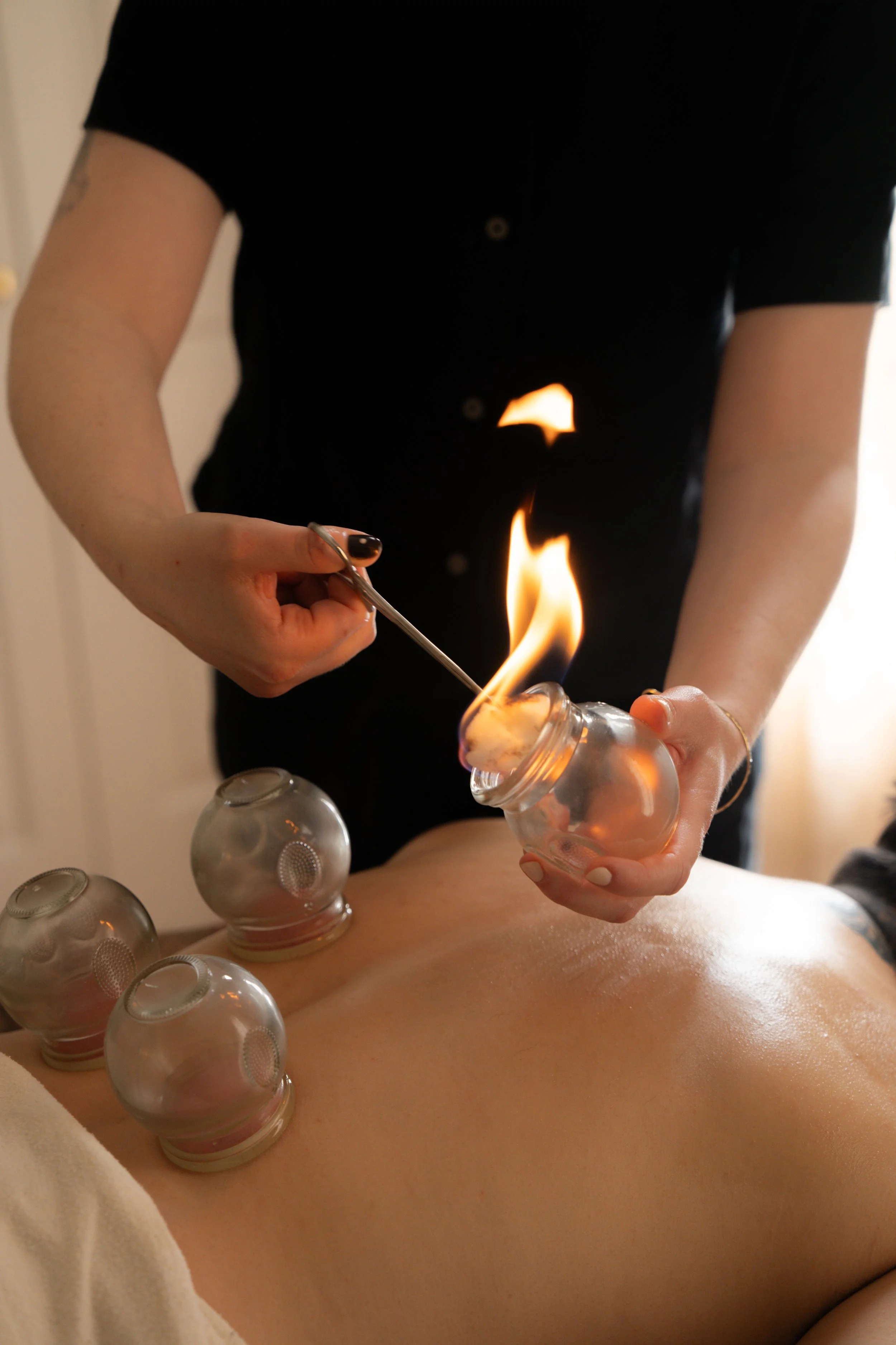 A person with black painted nails holding a lit match over a small bottle, with several empty bottles arranged on a person's bare leg.