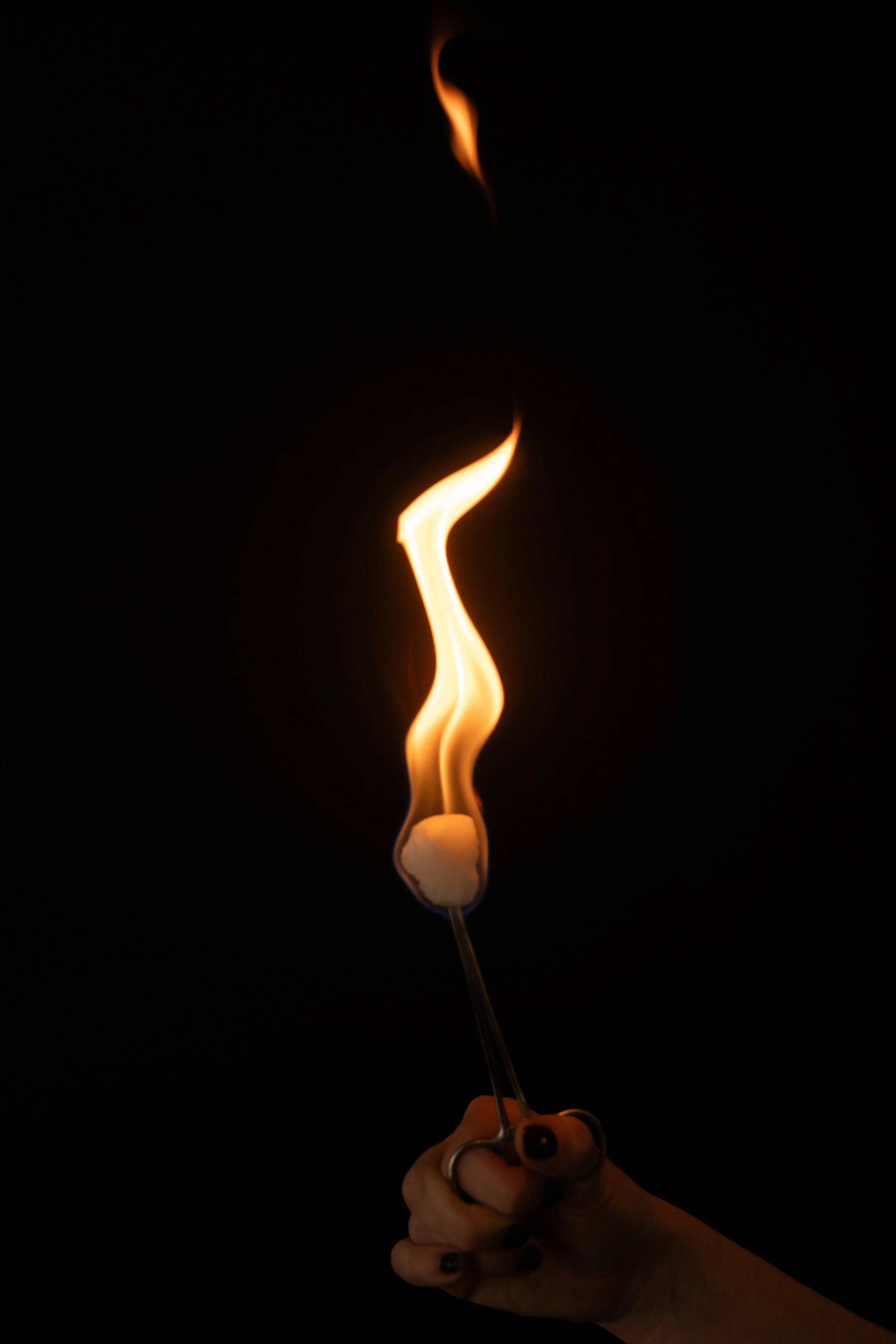 A person's hand holding a long cotton ball cupping instrument with a flame burning and rising into the air against a black background.