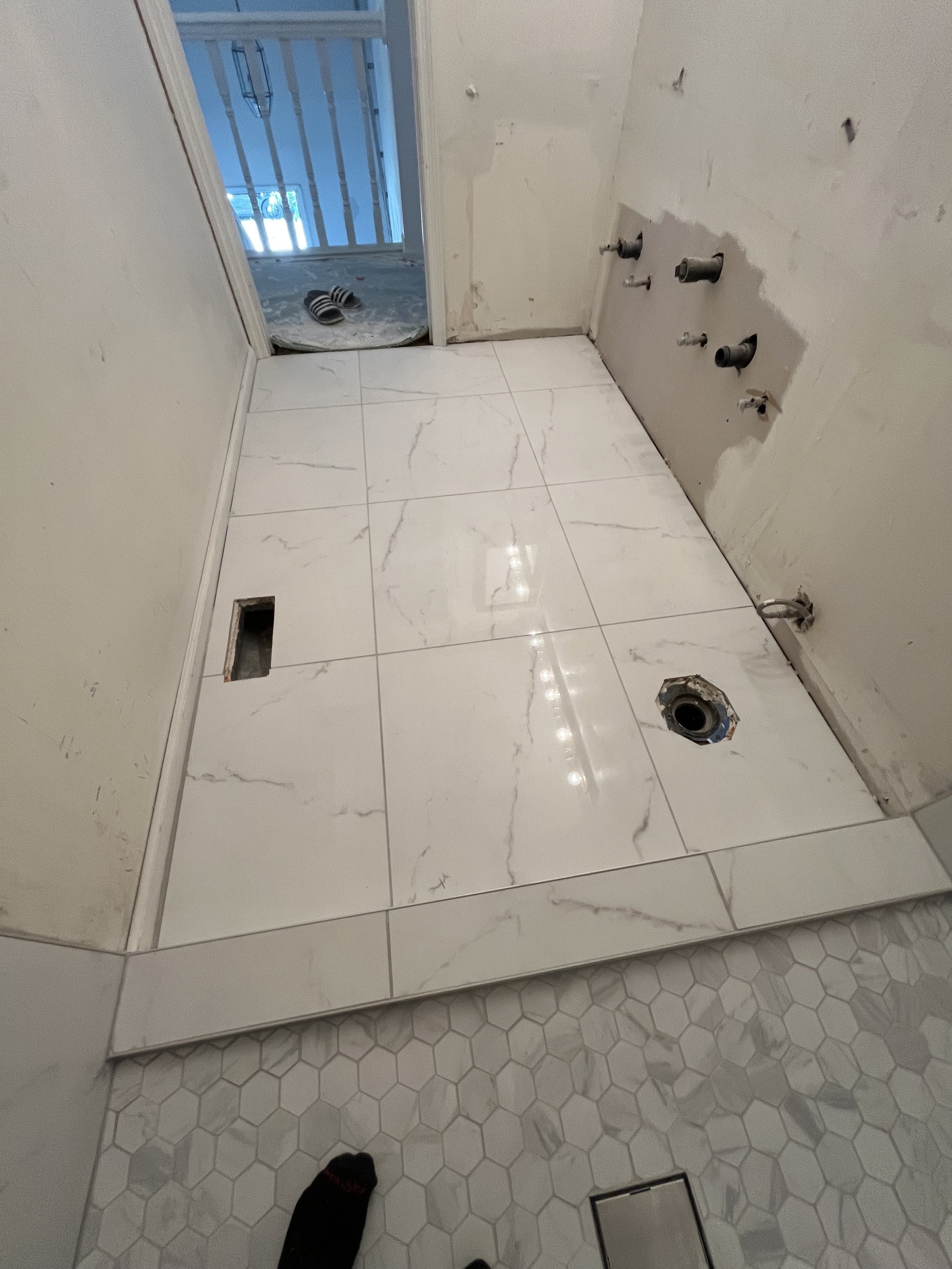 A bathroom under renovation with white marble floor tiles, visible plumbing pipes, an electrical outlet box, and a doorway leading to a balcony with slippers on the floor.