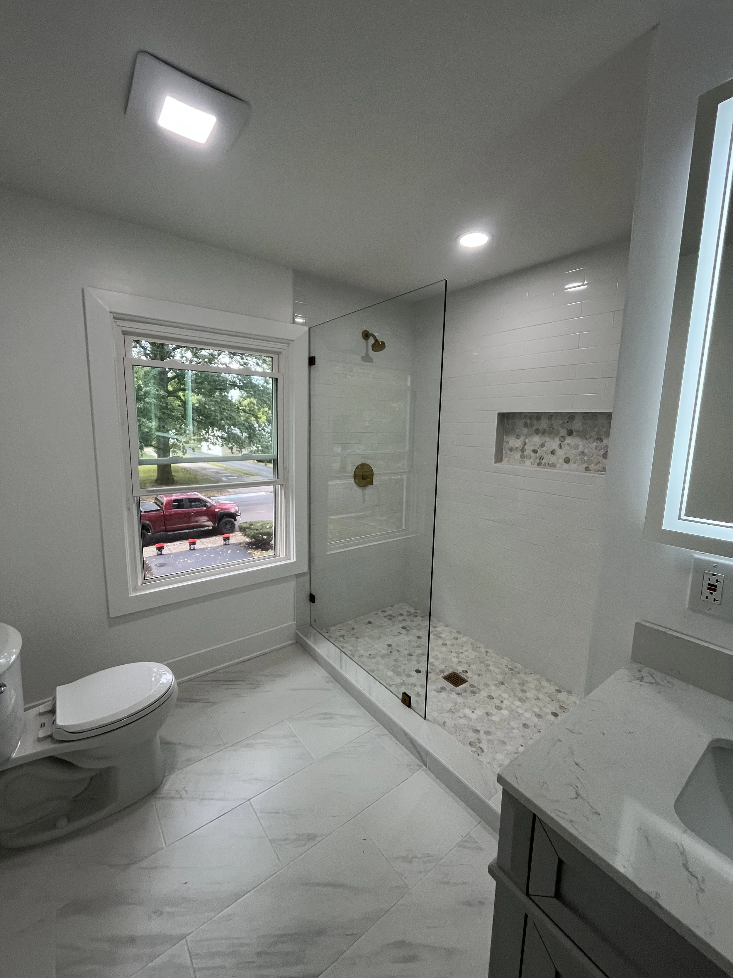Modern bathroom with white marble tiles, a toilet, a glass-enclosed shower with pebble floor, and a marble countertop with a sink. A window shows a view of a red truck and green trees outside.