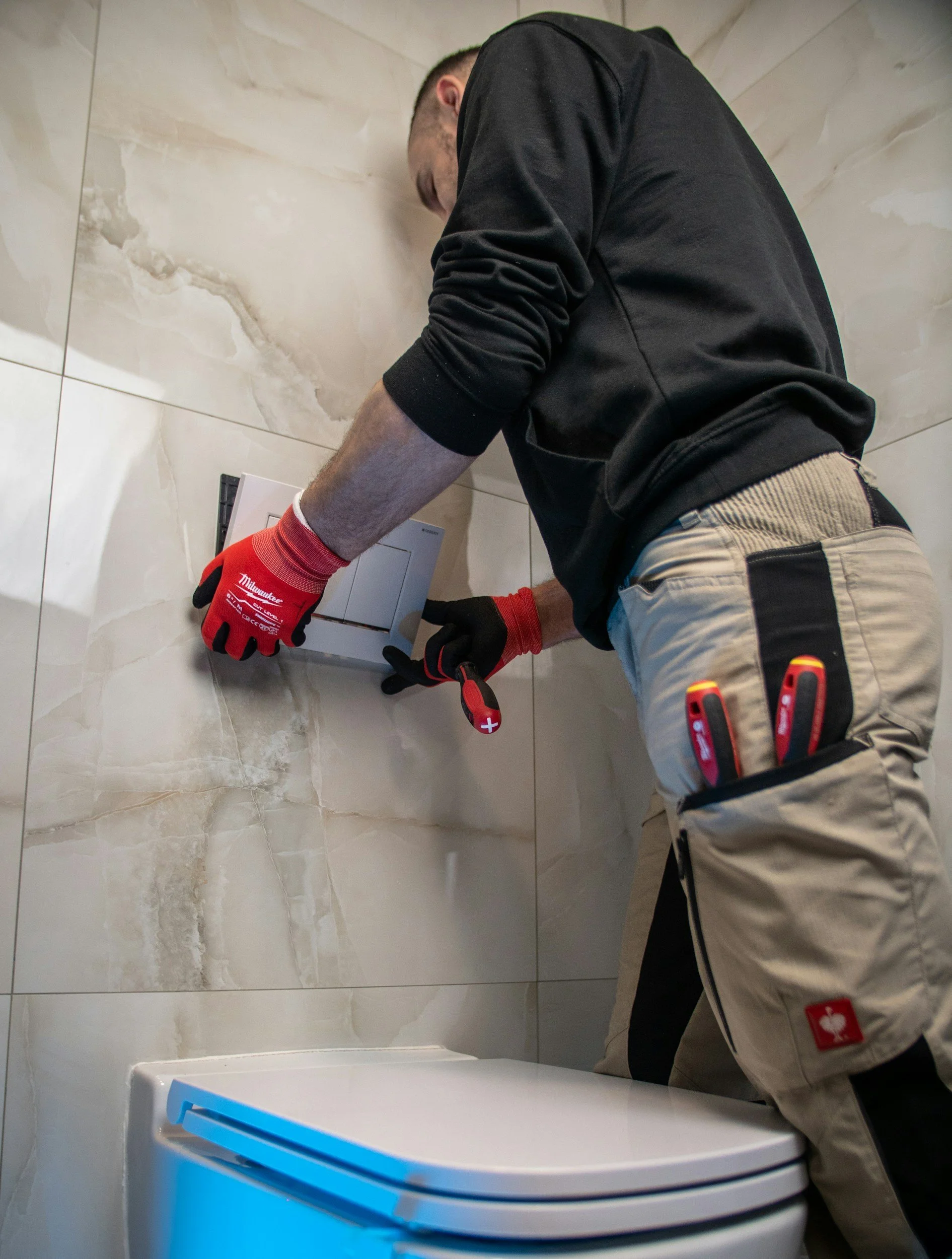 A worker is installing or repairing a light switch in a bathroom with beige marble walls, wearing red gloves and tools in his pocket.