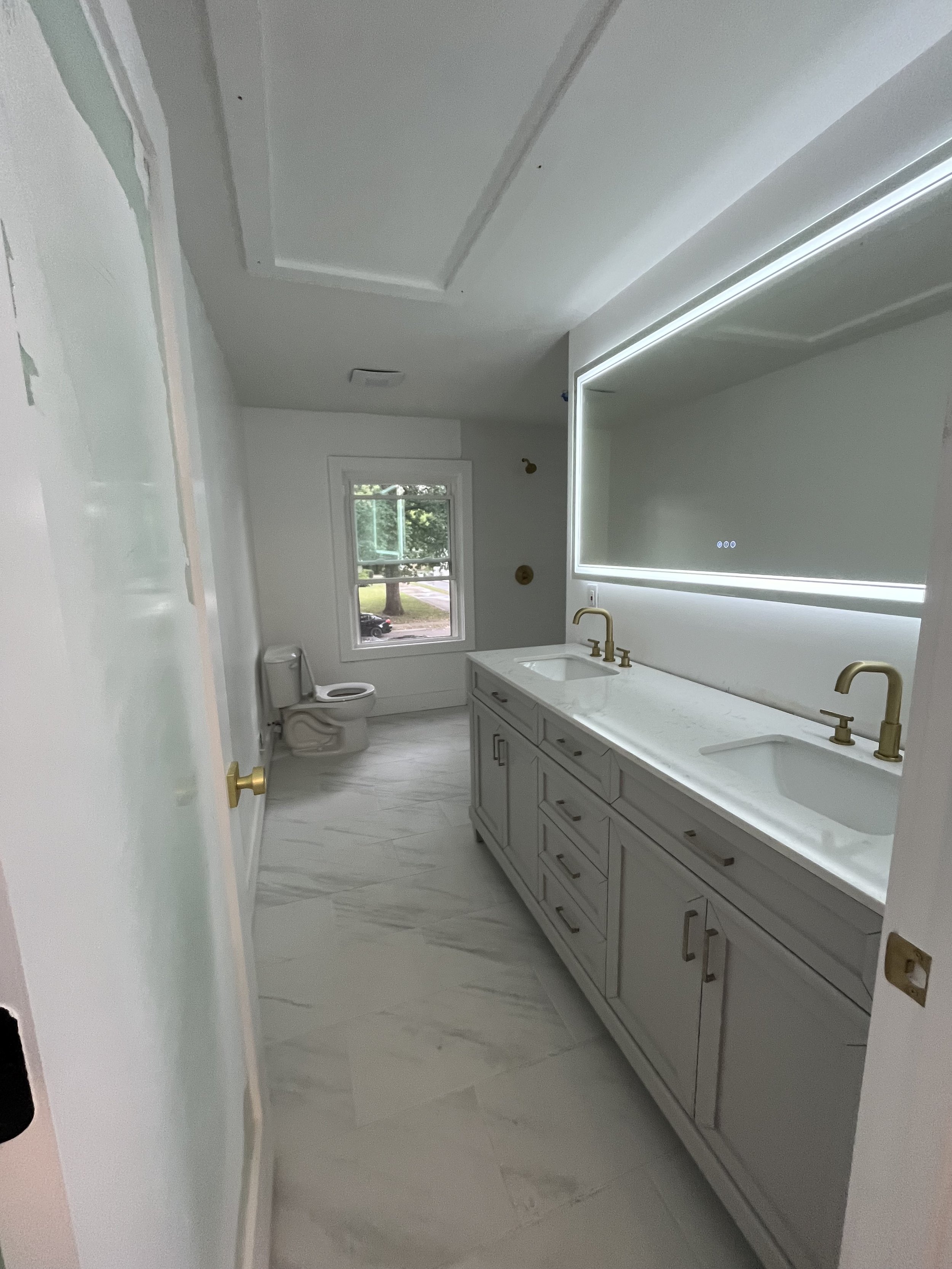 A modern bathroom under construction with a white double vanity with three drawers and four cabinets, gold faucets, and a large backlit mirror. There is a window showing an outdoor scene with trees and a street. A toilet is situated near the window. 