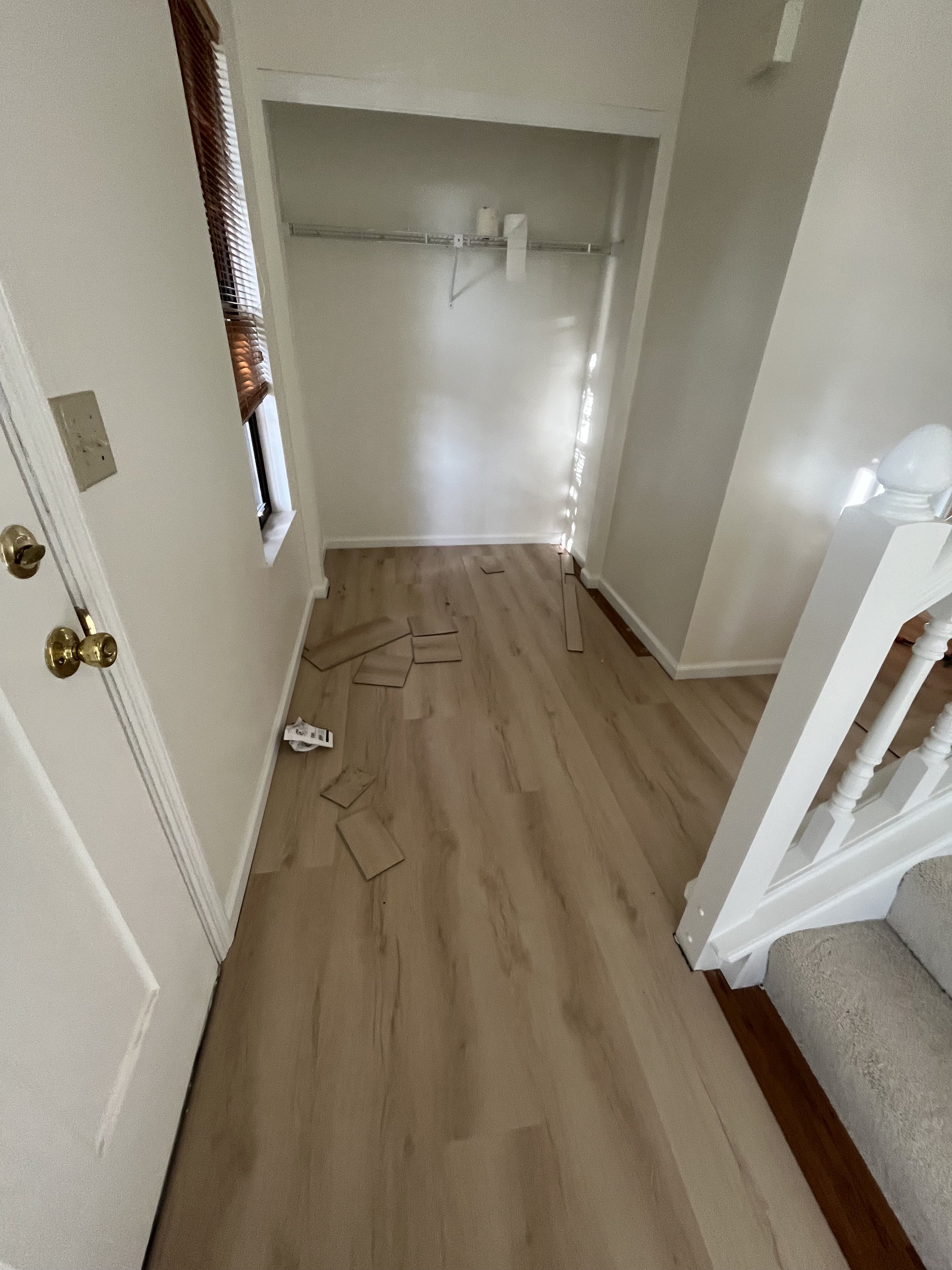 Empty hallway with a small closet containing a wire shelf and a few scattered boxes or papers on the wood floor, next to a staircase with carpeted steps and a white railing.
