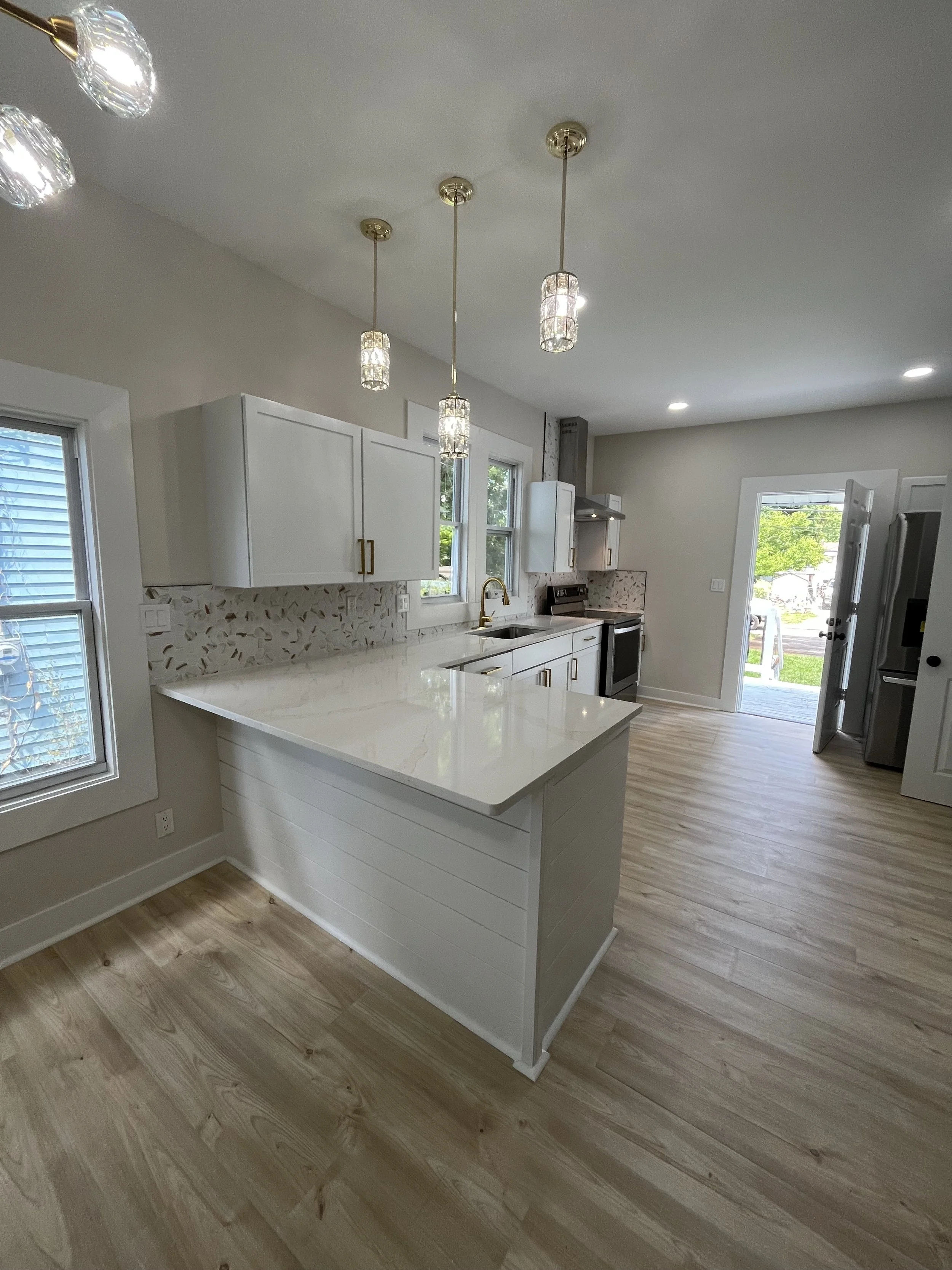 Bright, modern kitchen with white cabinets, a marble countertop island, pendant lights, hardwood floors, and natural light from windows and a door leading outside.