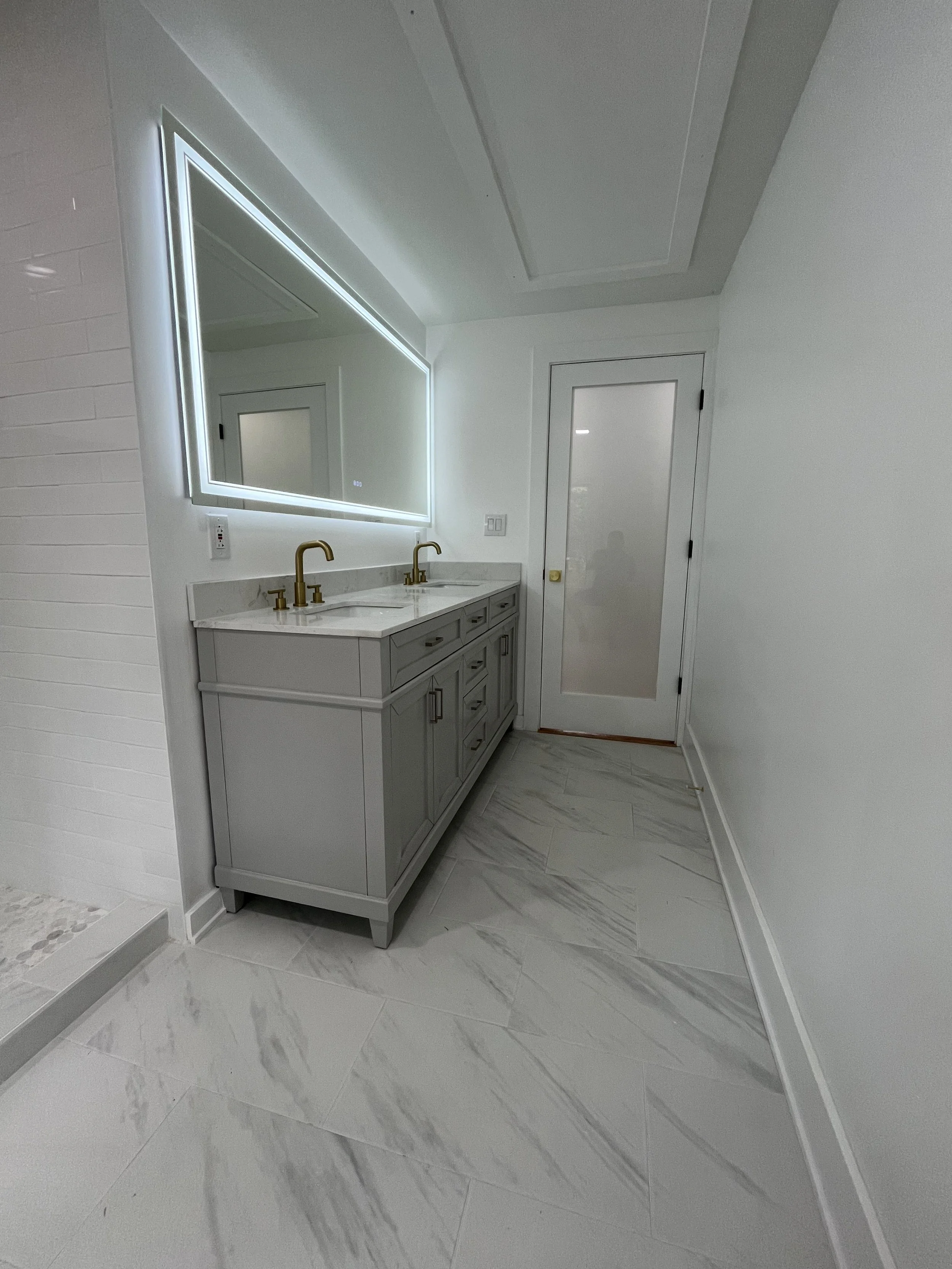 Modern bathroom with double vanity, marble countertops, gold fixtures, illuminated mirror, frosted glass door, and marble tiled floor.