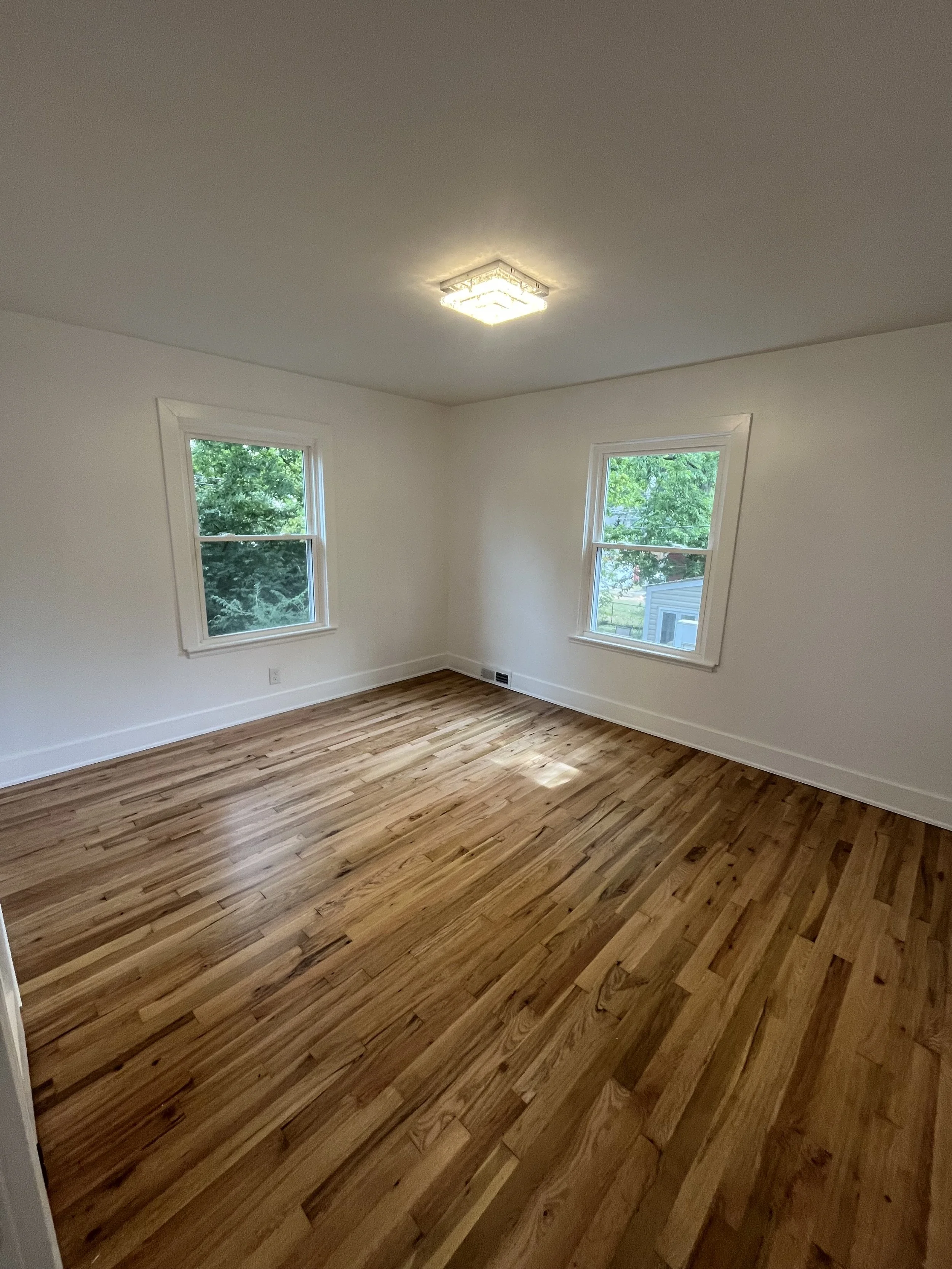 Empty room with hardwood floors, white walls, two windows with view of green trees, and ceiling light fixture.