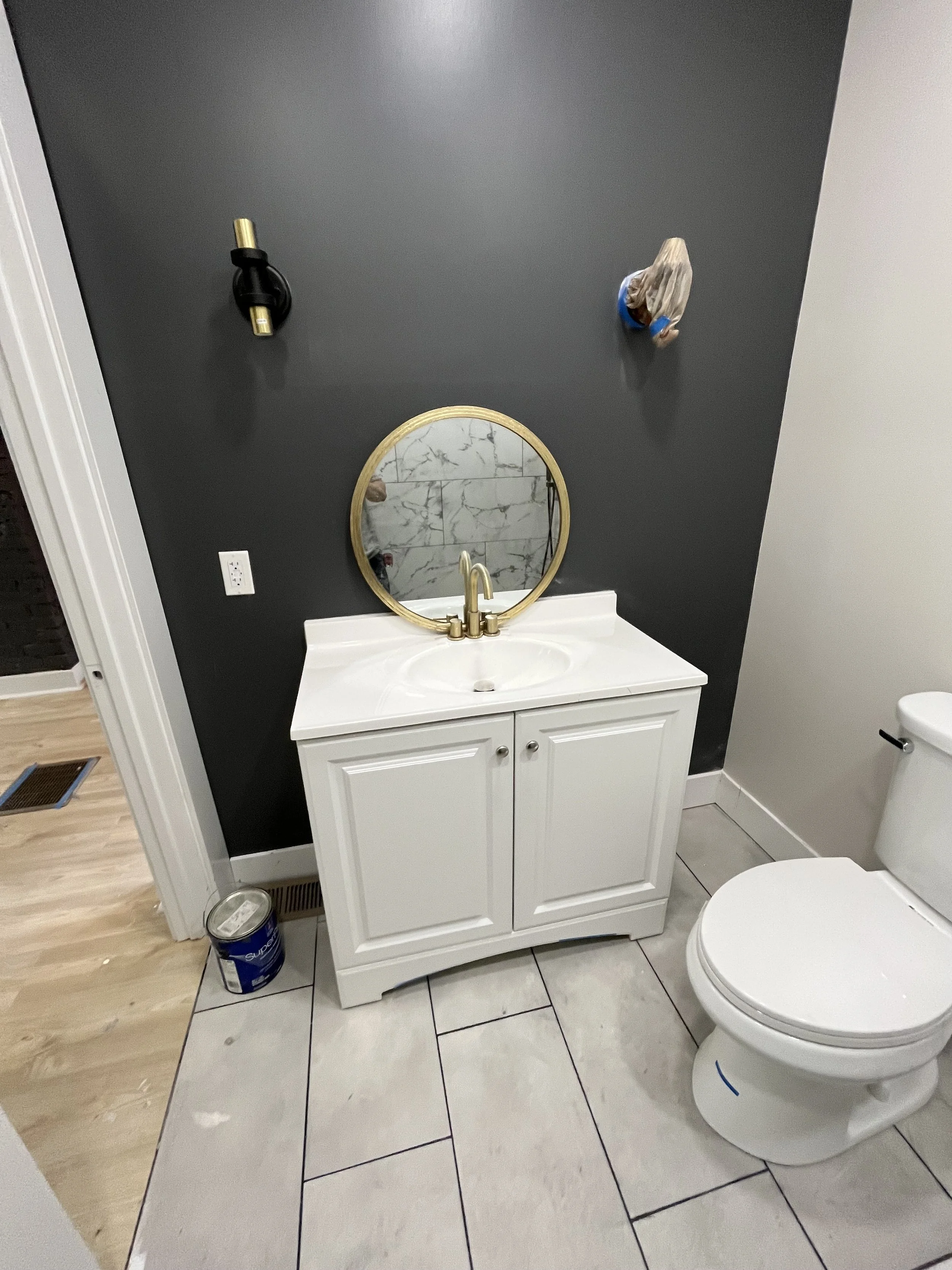 Small bathroom with a white vanity sink, a round mirror with a gold frame, a dark gray accent wall, two wall fixtures, and a toilet. The floor has large light-colored tiles.